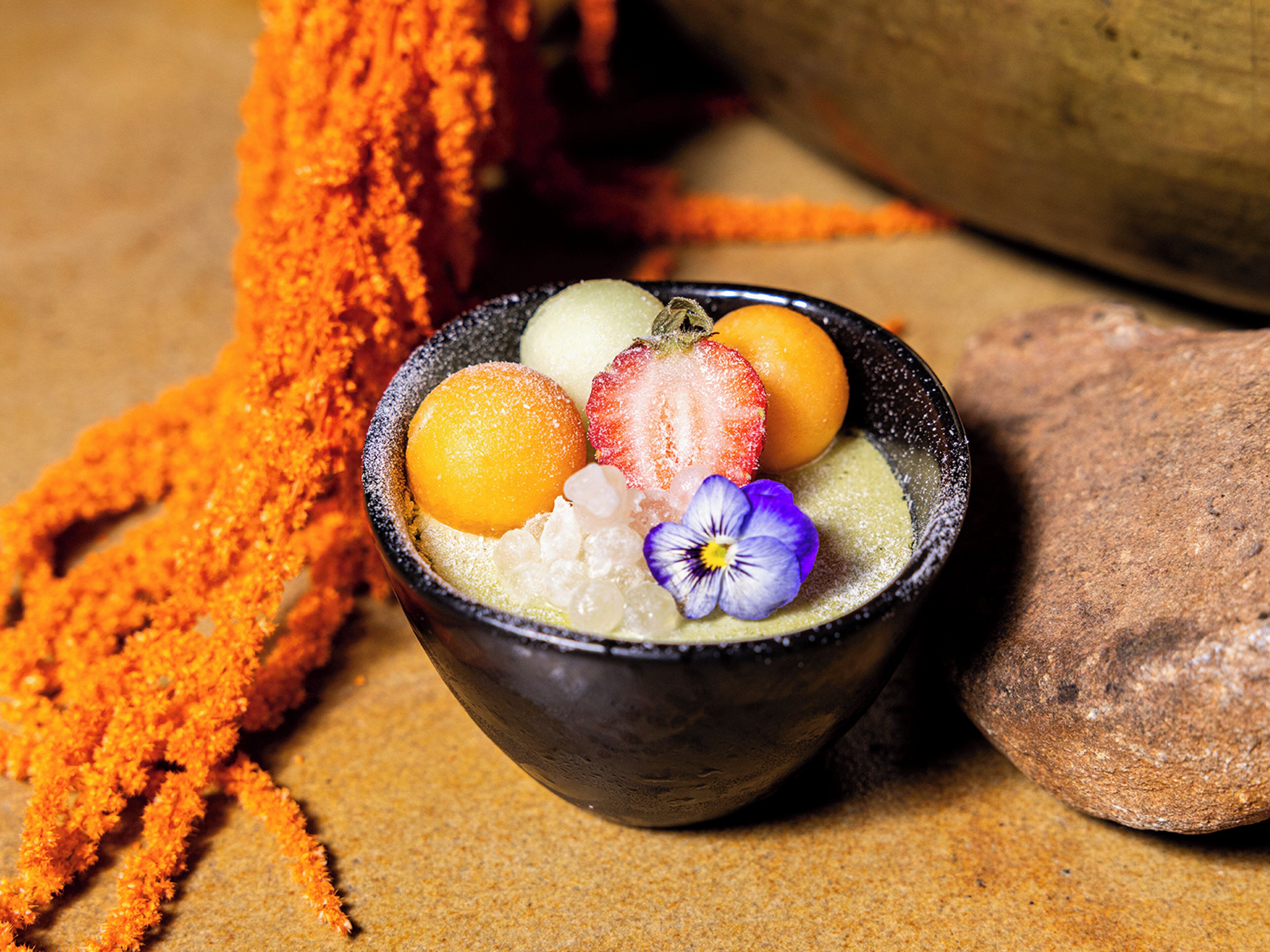 A bowl of dessert with fruits and a flower, placed on a textured surface with a rock and orange powder nearby.