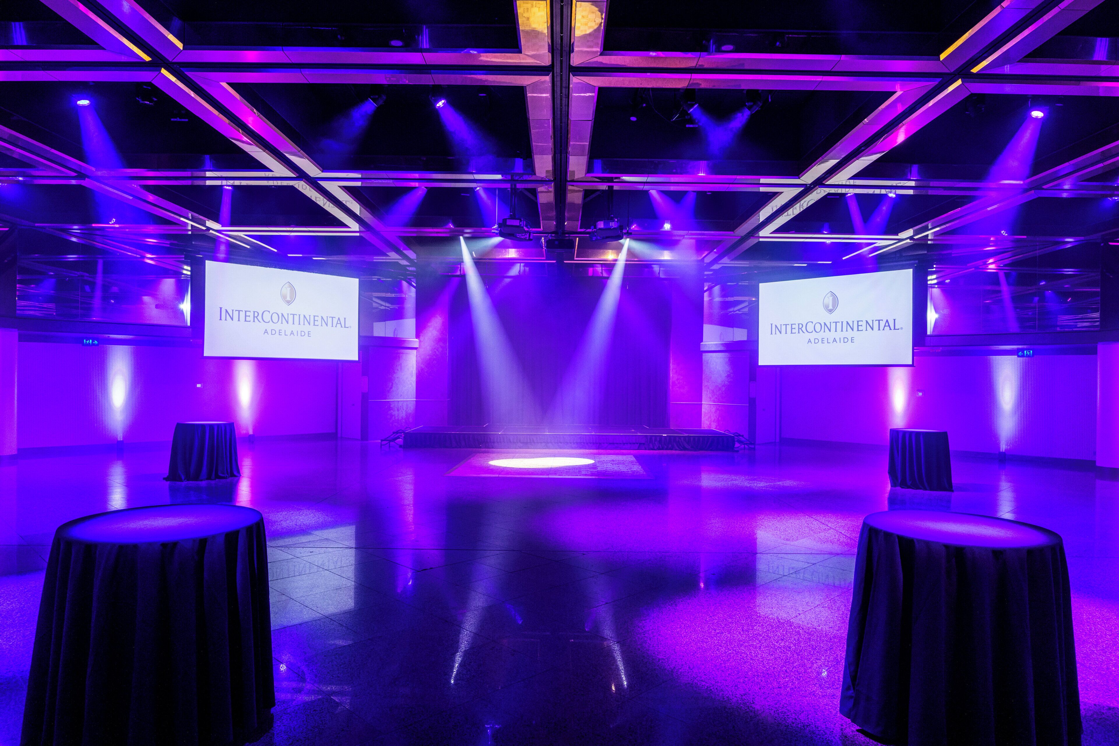 Een congreszaal met een podium, twee grote schermen waarop 'Intercontinental Adelaide' staat, en een gepolijste vloer.