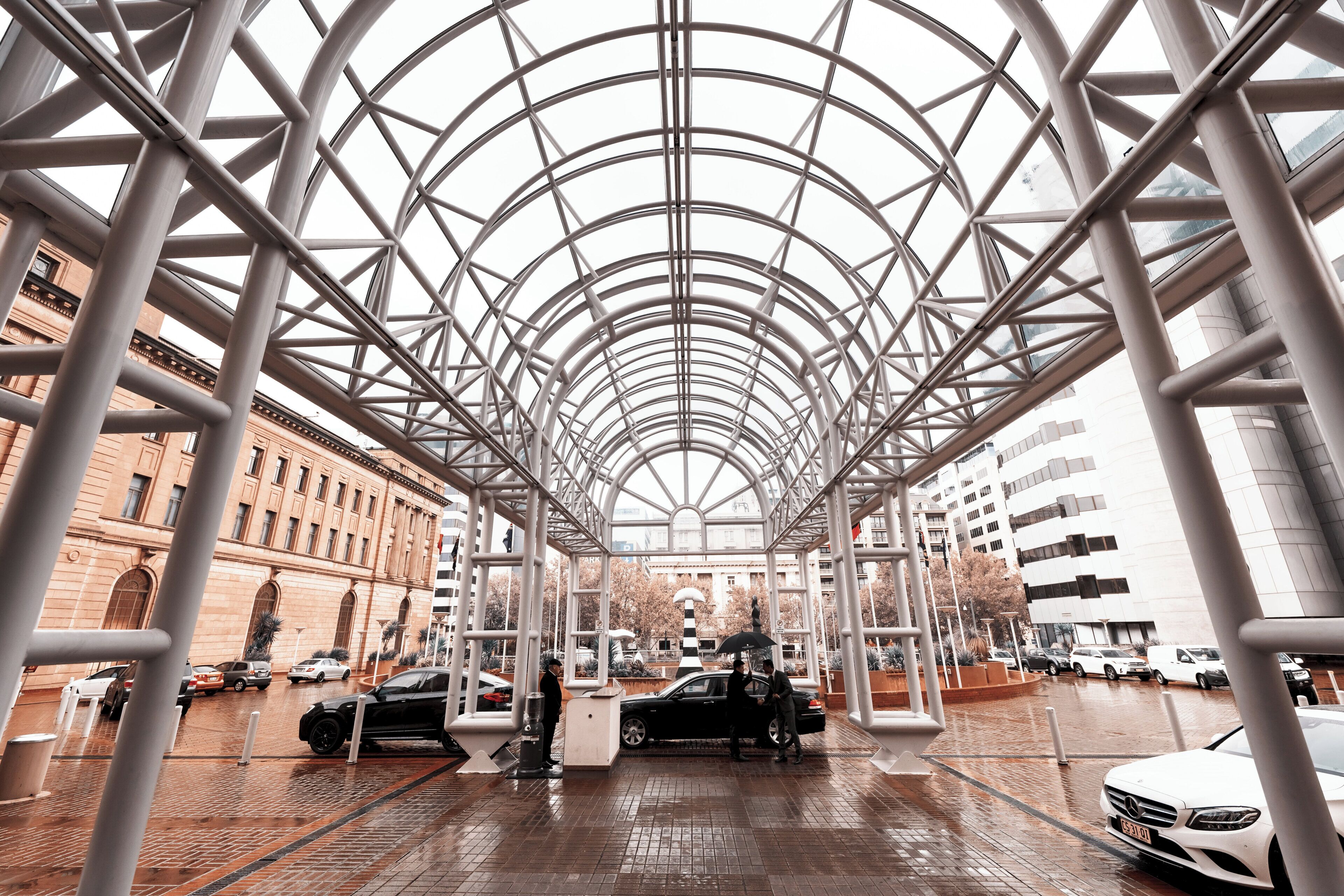 A covered pedestrian walkway with a glass ceiling, surrounded by buildings, and cars parked nearby.
