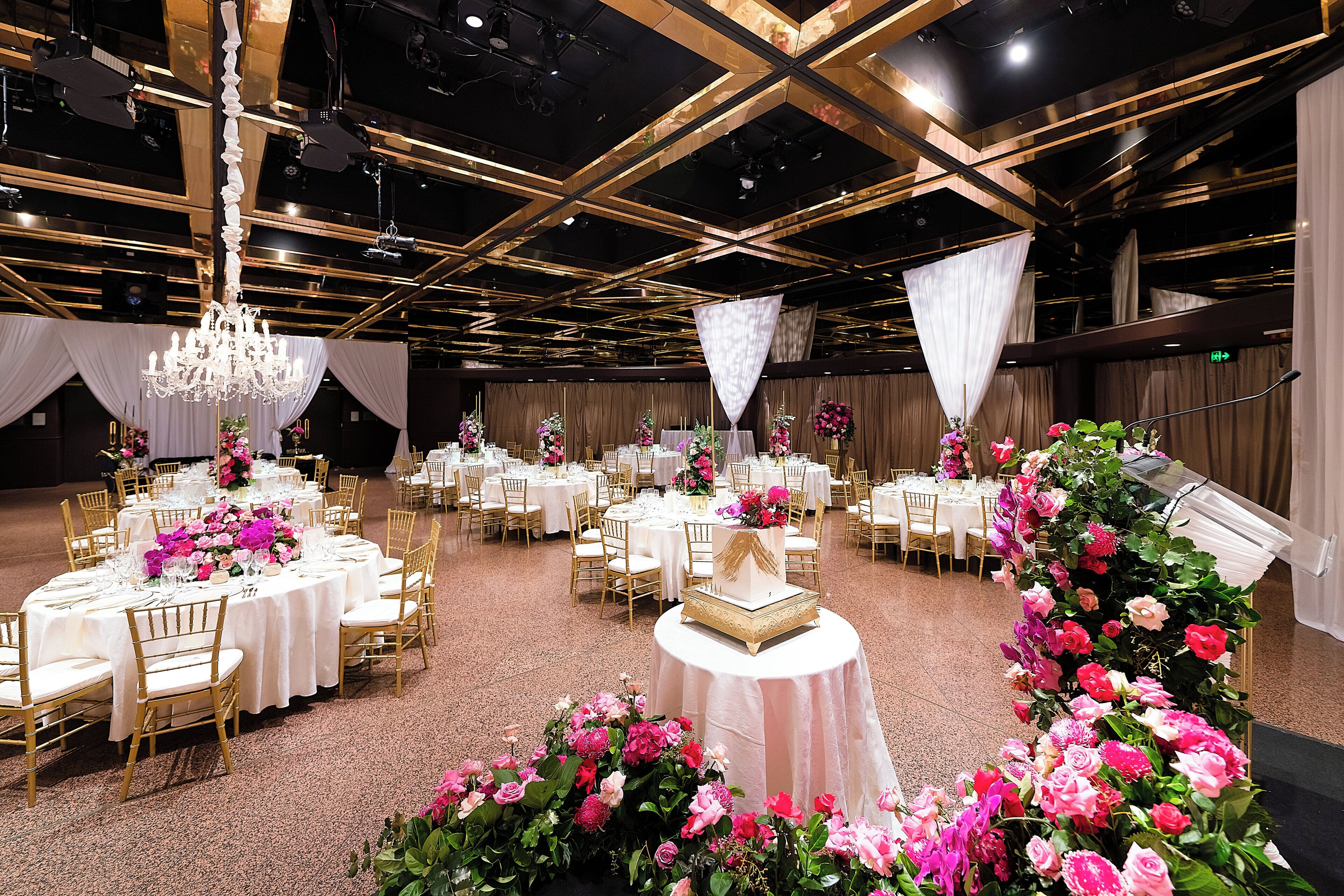 A banquet hall with round tables, golden chairs, and floral centerpieces.