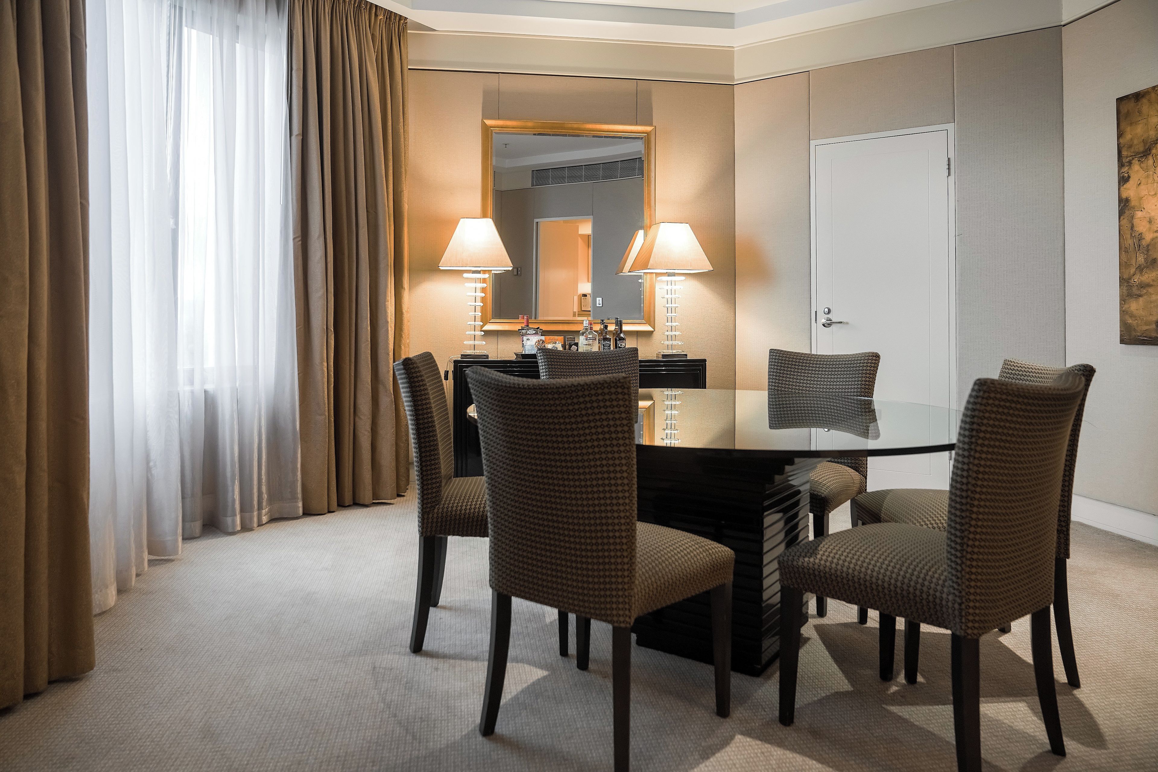 A hotel room with a round table, four chairs, two lamps, a mirror, and a door.