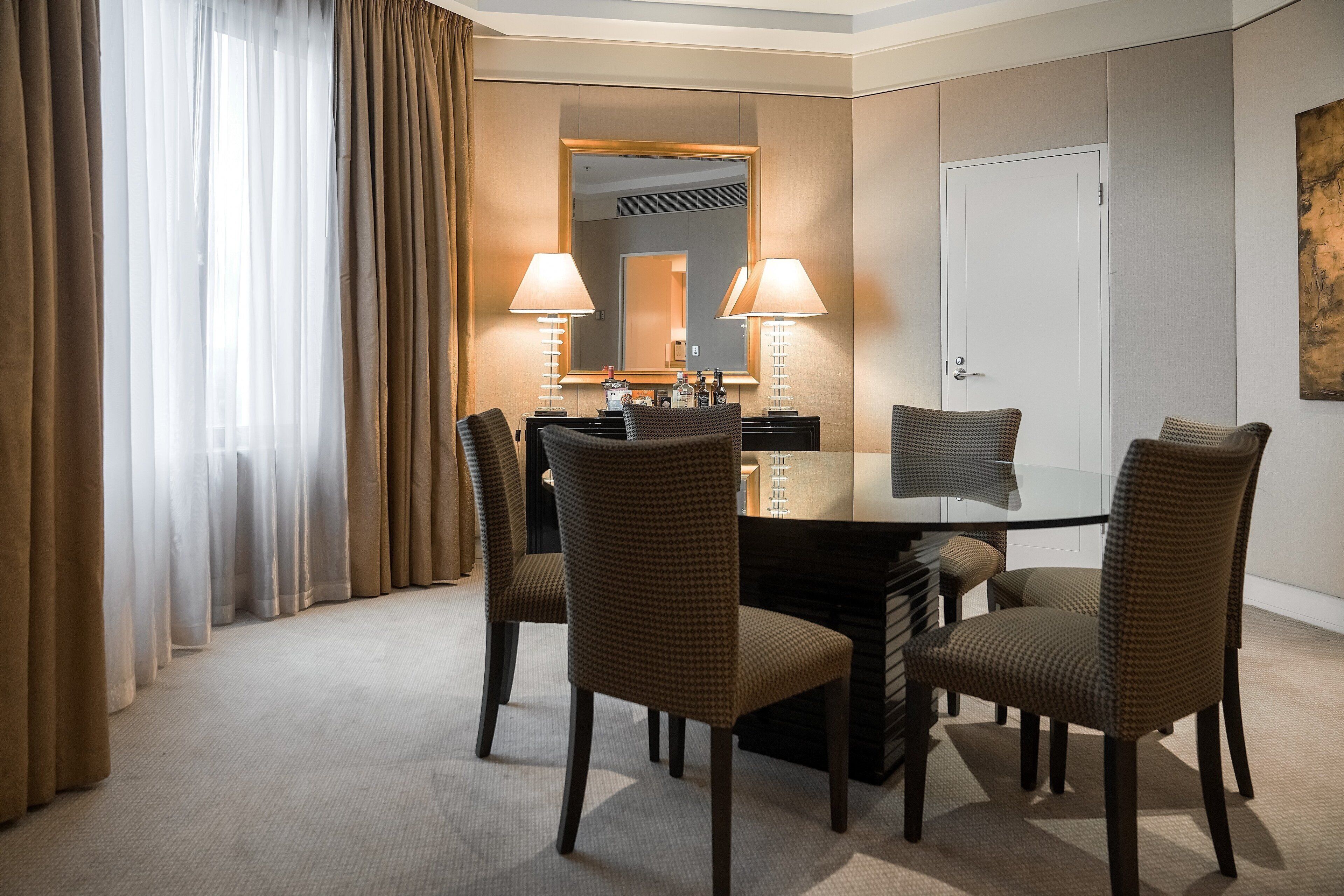 Habitación de hotel con una mesa redonda, cuatro sillas, dos lámparas, un espejo, una puerta y un cuadro en la pared.