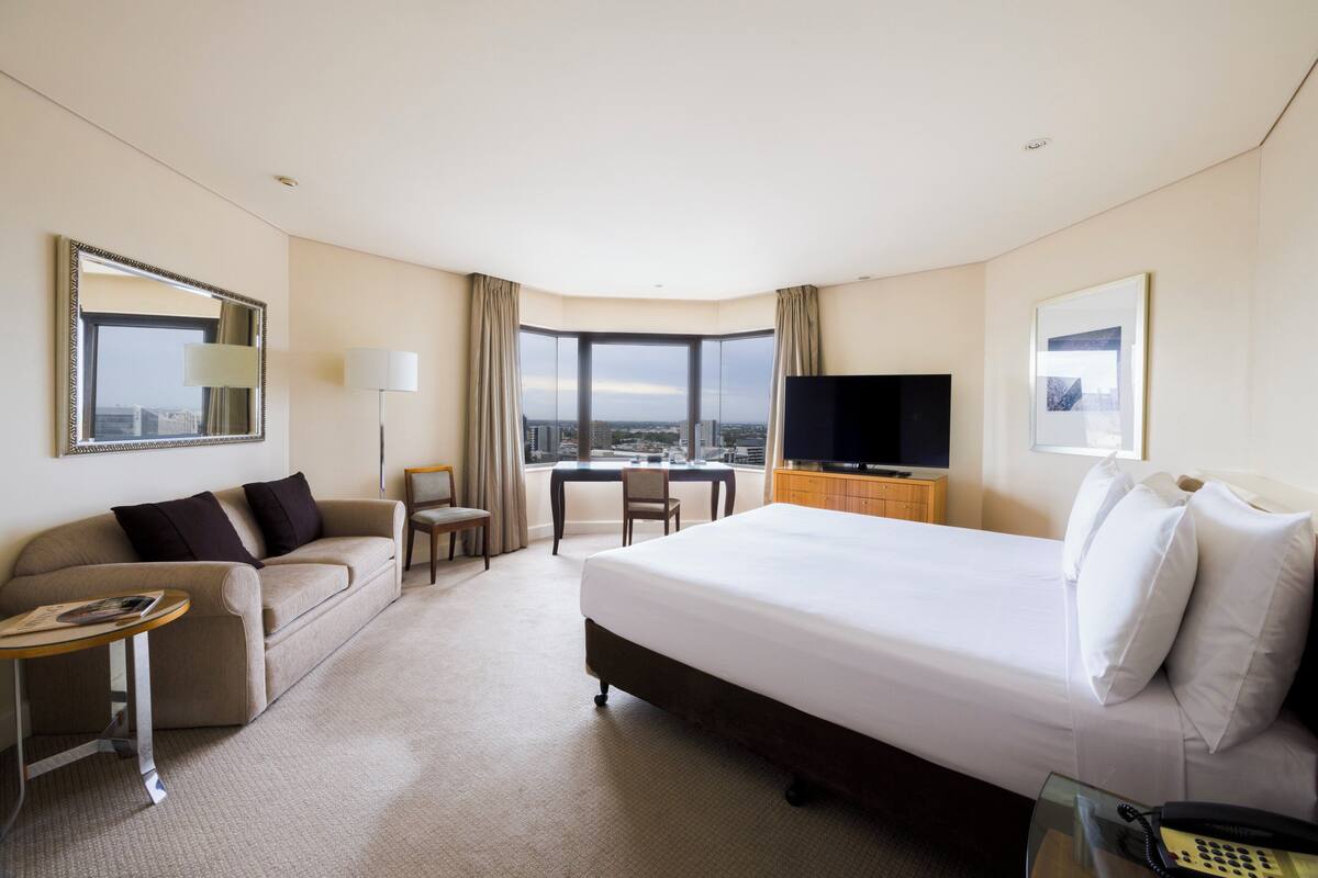 Habitación de hotel con una cama grande, un sofá, un escritorio, un televisor y vistas de la ciudad.