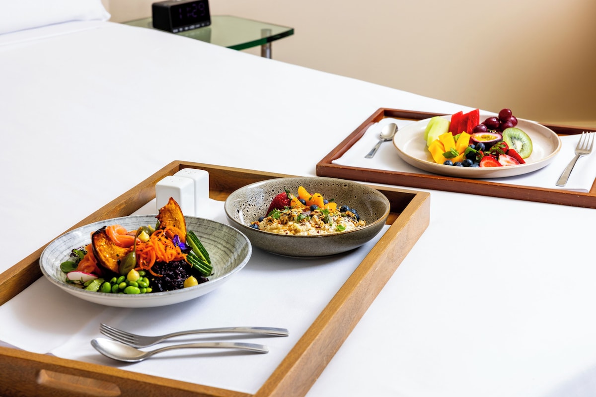 A breakfast tray with two bowls of food, a spoon, and a fork on a bed.