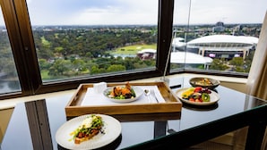 A dining table with plates of food and a tray, set against a window with a view of a stadium and greenery. - InterContinental Adelaide by IHG (Adelaide)