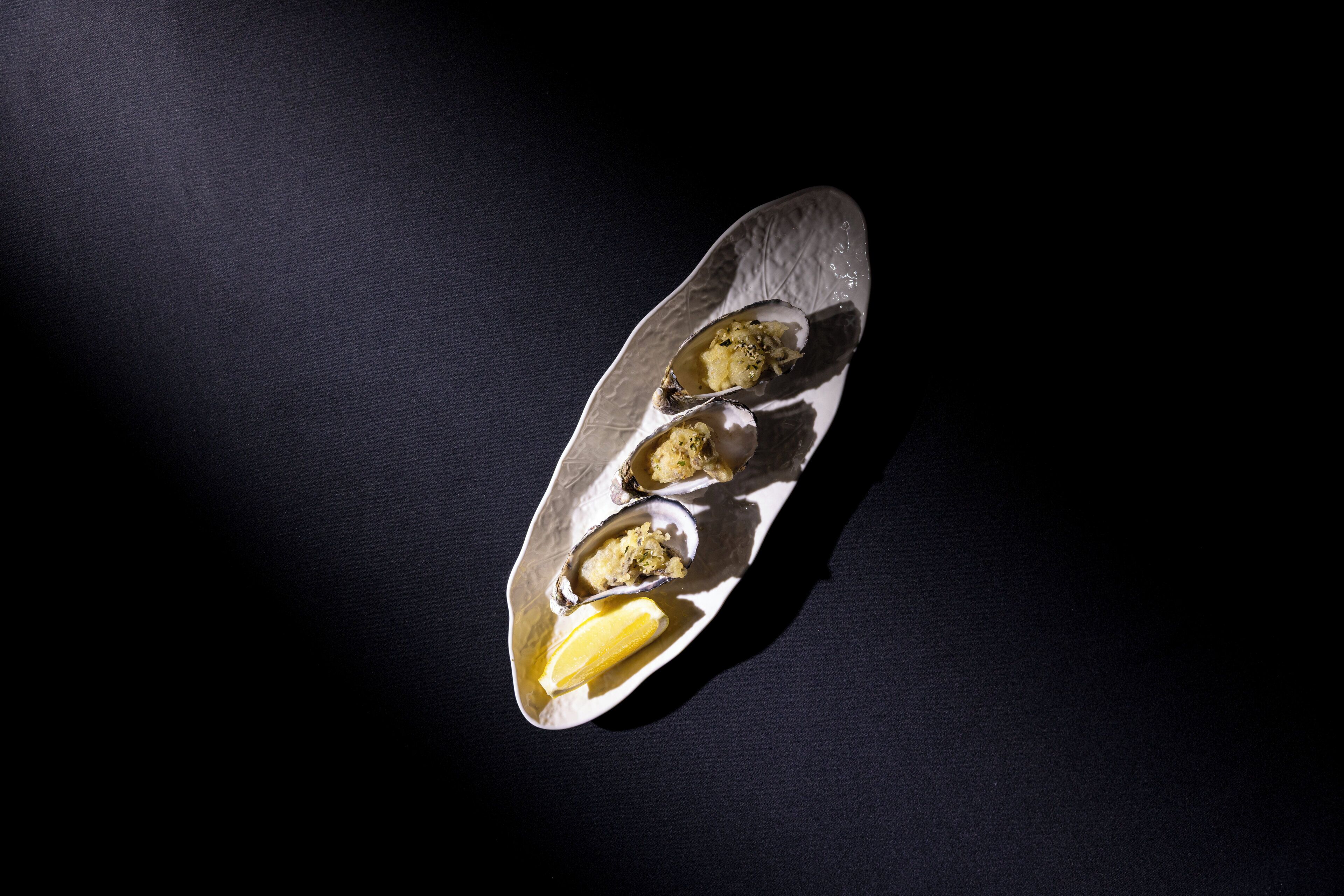 A plate with three oysters and a lemon wedge on a dark surface.