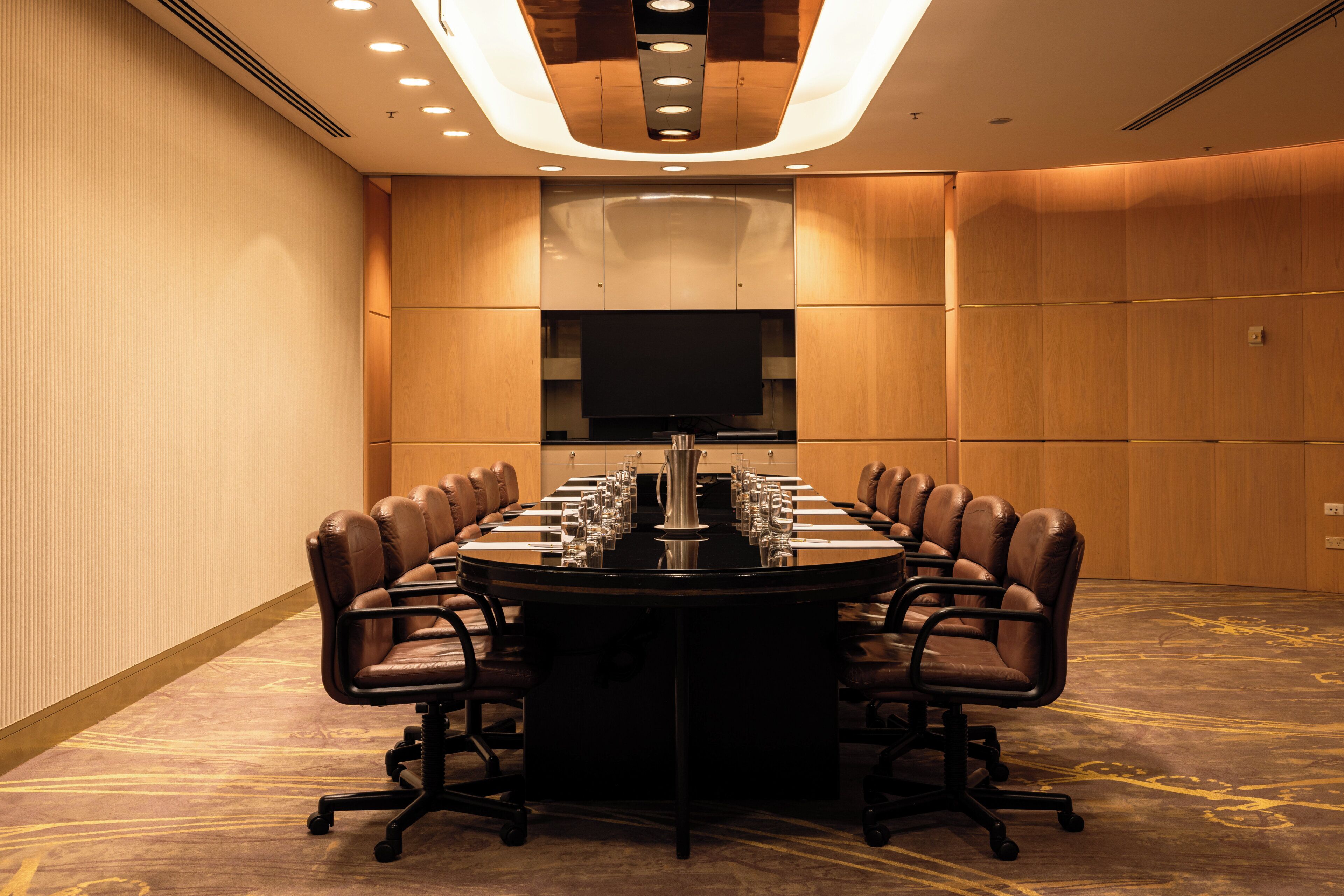 A conference room with a long table, leather chairs, a flat-screen TV, and recessed ceiling lighting.
