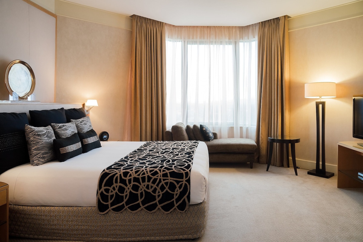 A hotel room with a large bed, a sitting area, a television, and a window with curtains.