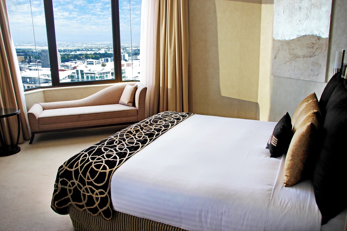 A hotel room with a large bed, a sofa, and a view of the cityscape through the window.