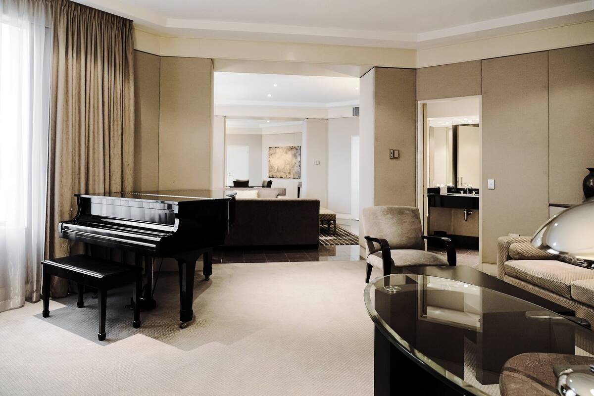 Una habitación de hotel moderna con un piano, una mesa de centro de vidrio, un sofá y un espejo grande.