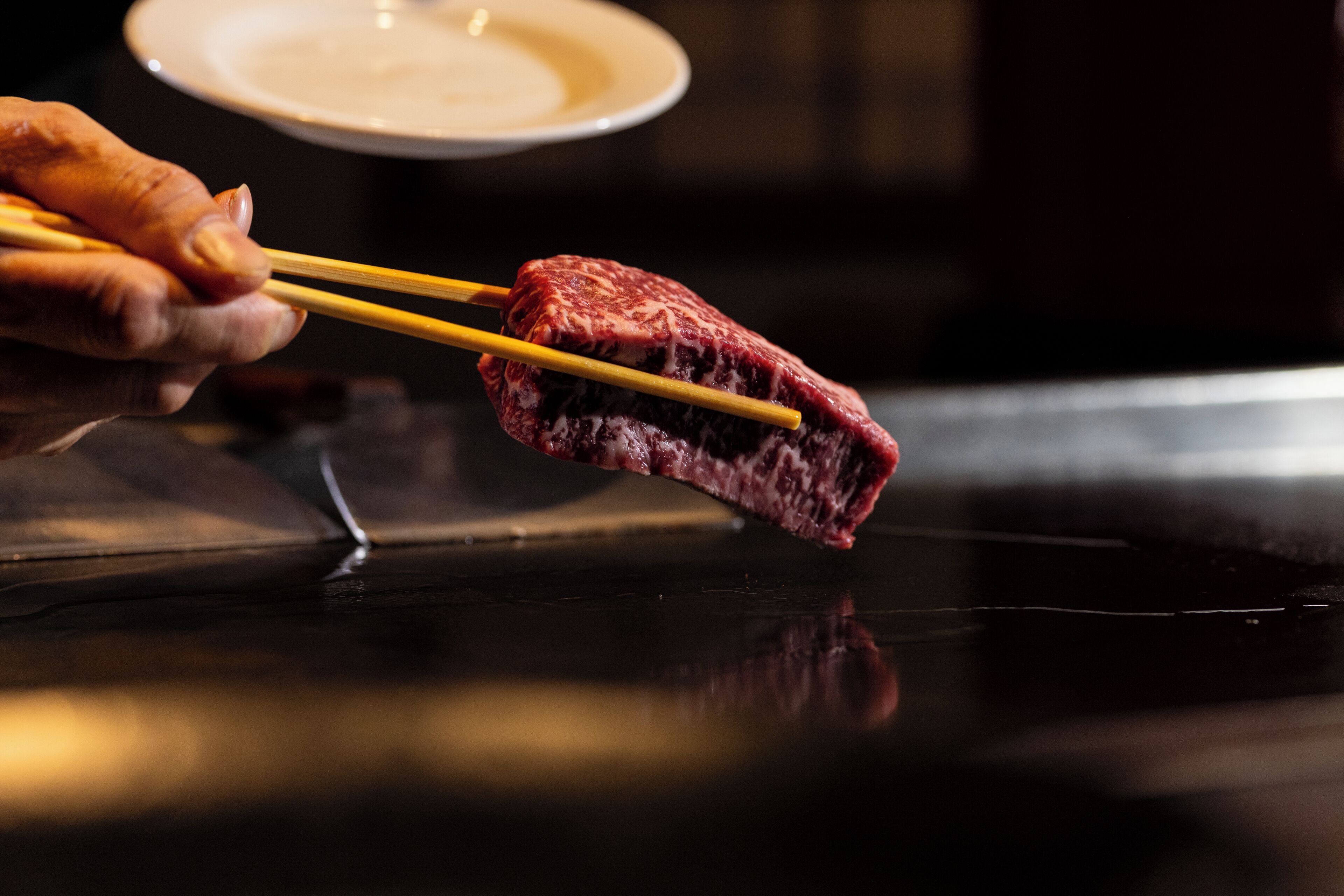 A hand holding a piece of raw meat on a pair of chopsticks over a grill.