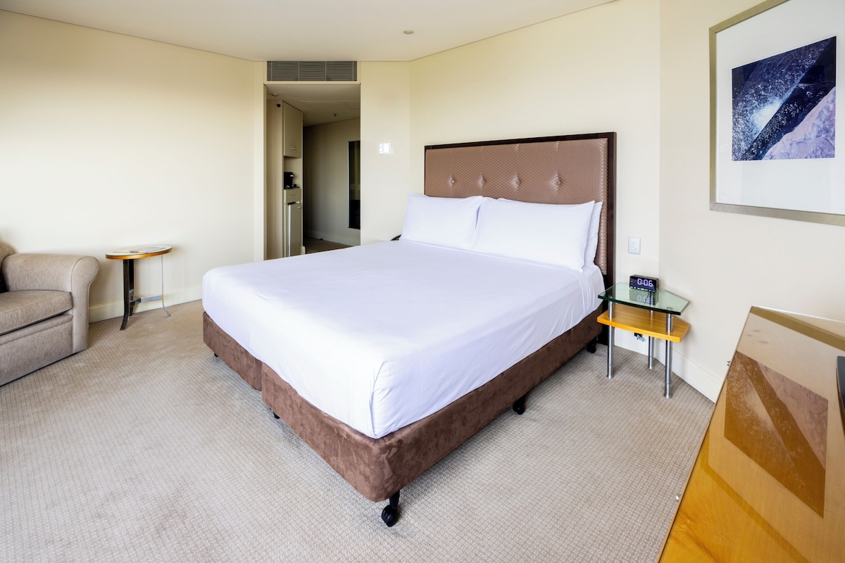 A hotel room with a large bed, a small table, a chair, and a painting on the wall.