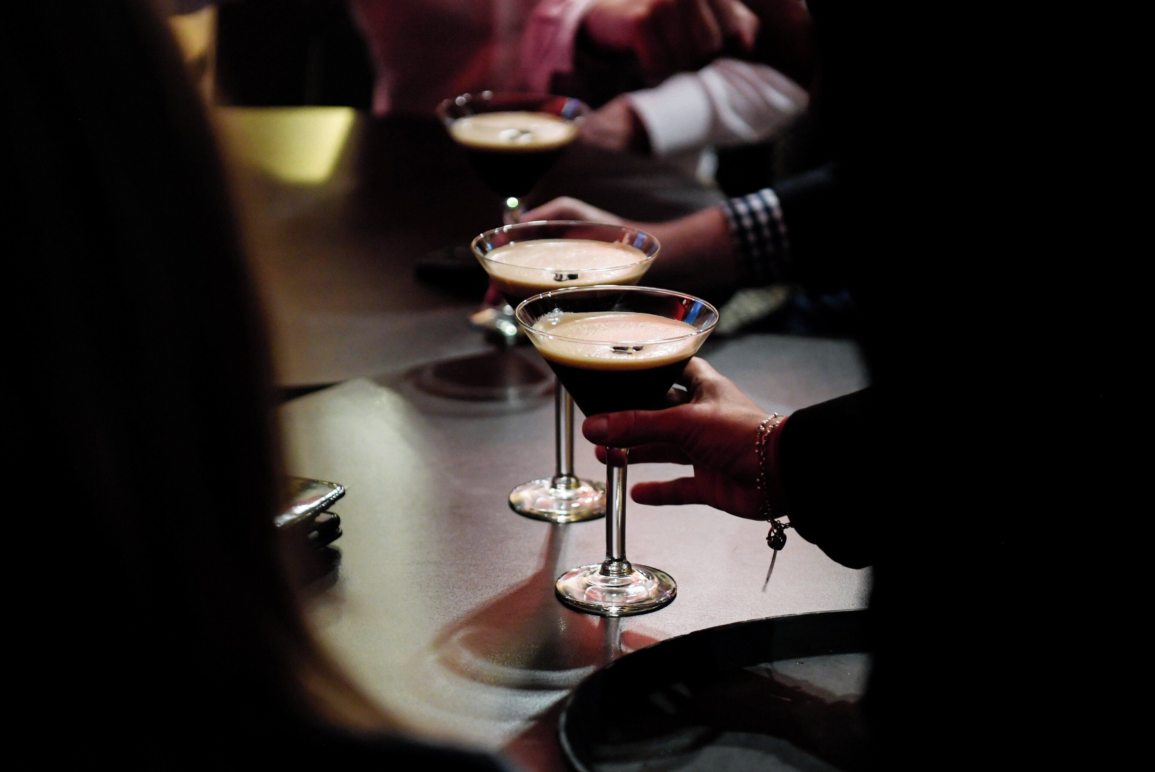 A bar setting with glasses of a dark beverage, likely coffee or a cocktail, with a focus on the drinks and hands holding them.