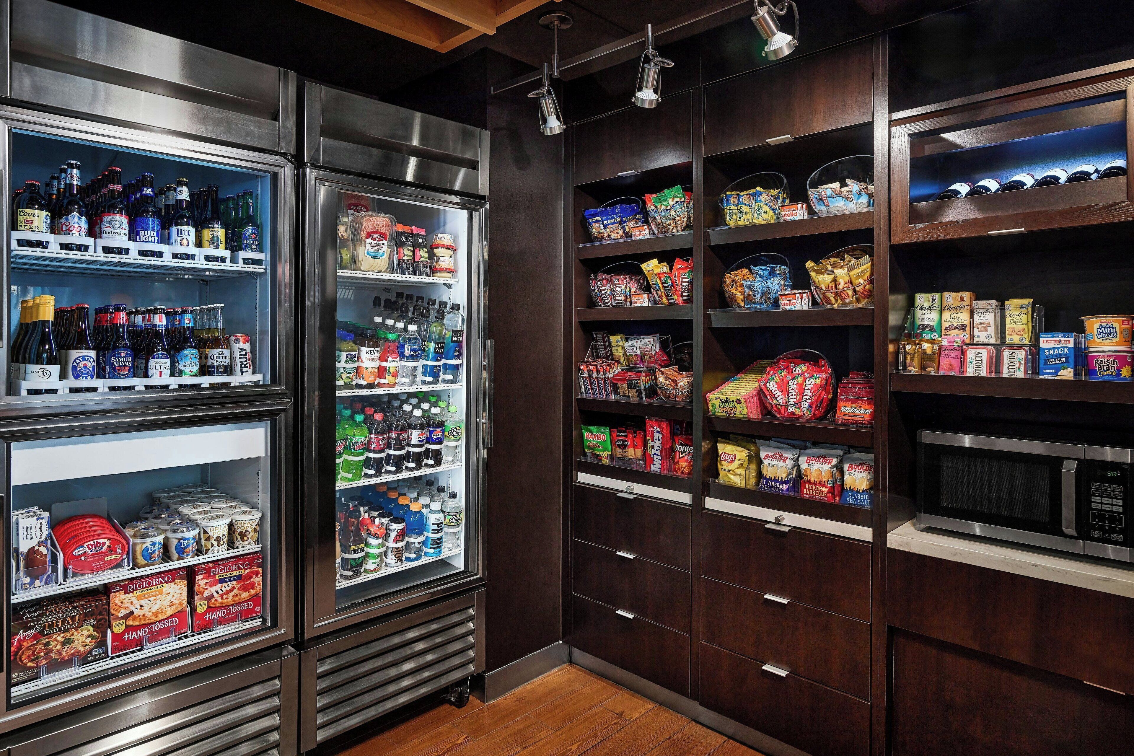 Un refrigerador bien surtido con bebidas, estantes con aperitivos y un microondas en un entorno comercial.