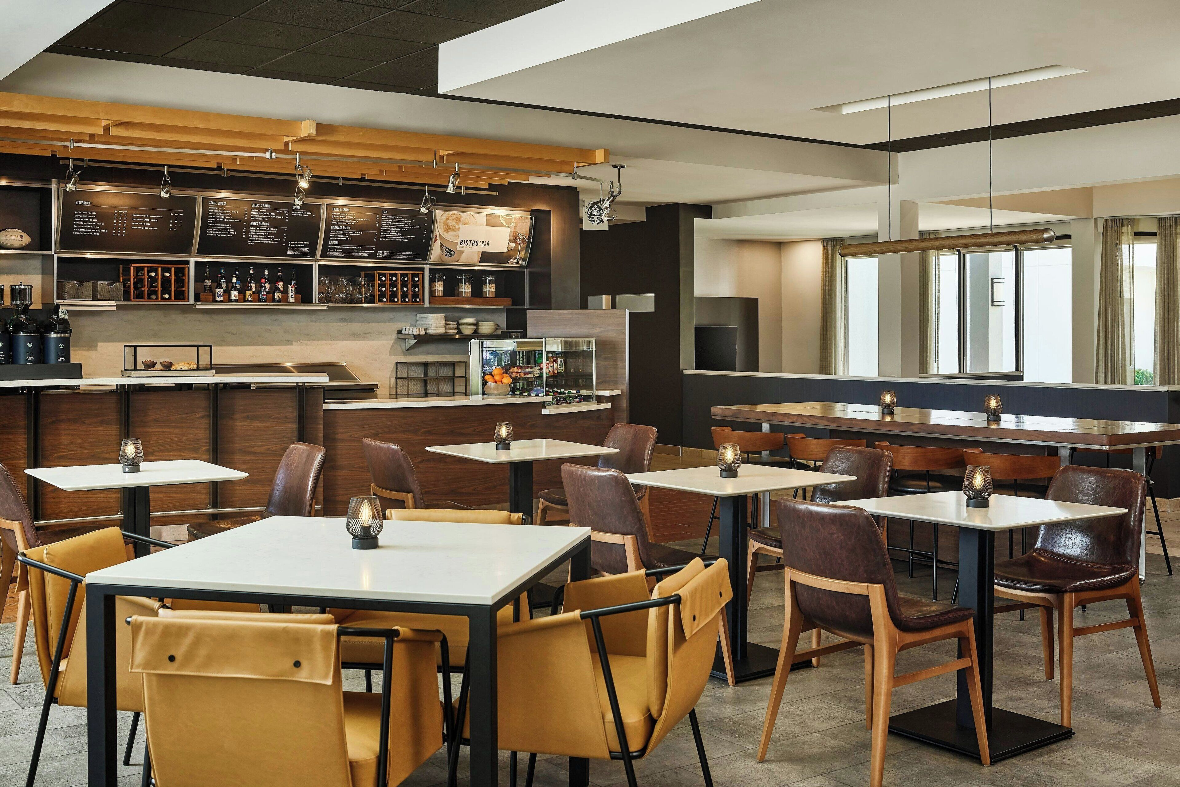 A modern restaurant interior with multiple tables and chairs, a bar area with shelves of bottles, and a large window providing natural light.