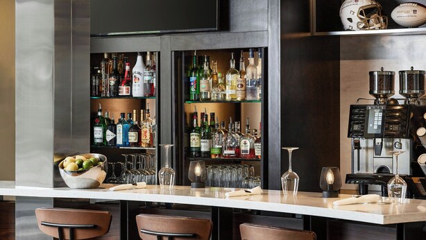 A bar area with a long counter, multiple shelves of liquor bottles, glasses, and a bowl of fruit. There are three bar stools and a television mounted on the wall. - Courtyard by Marriott Boulder (Boulder)