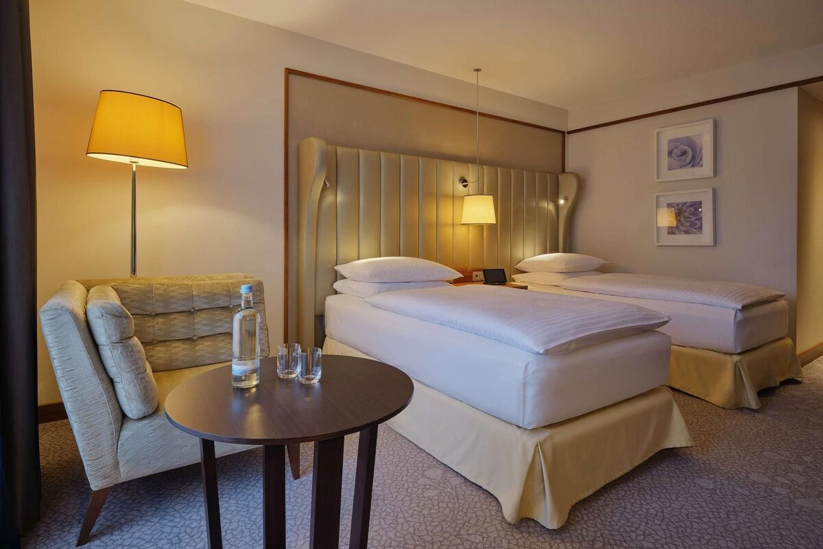 A hotel room with a bed, bedside tables, a chair, a lamp, and a small table with glasses and a bottle.