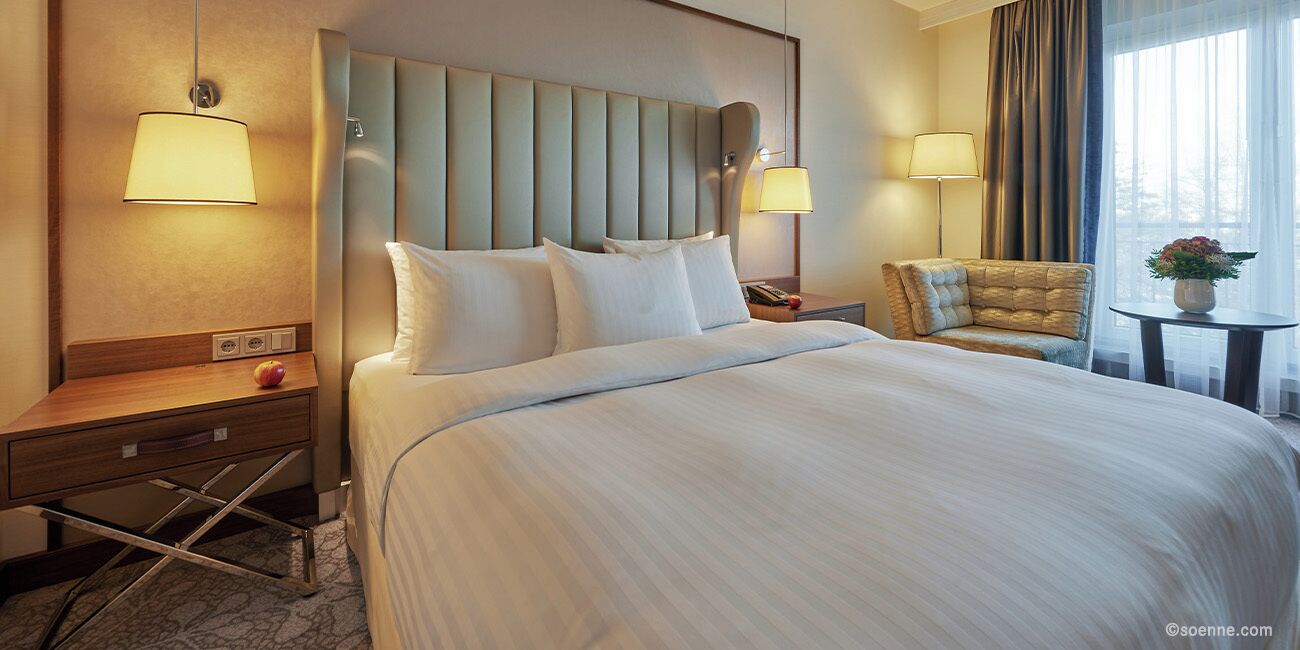 A hotel room with a large bed, two bedside lamps, a nightstand, a chair, and a small table with a vase.