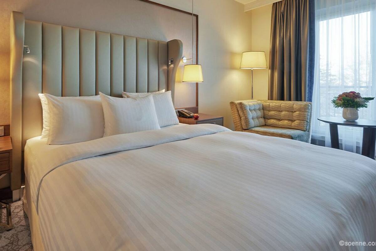 A hotel room with a large bed, two bedside lamps, a nightstand, a chair, and a small table with a vase.