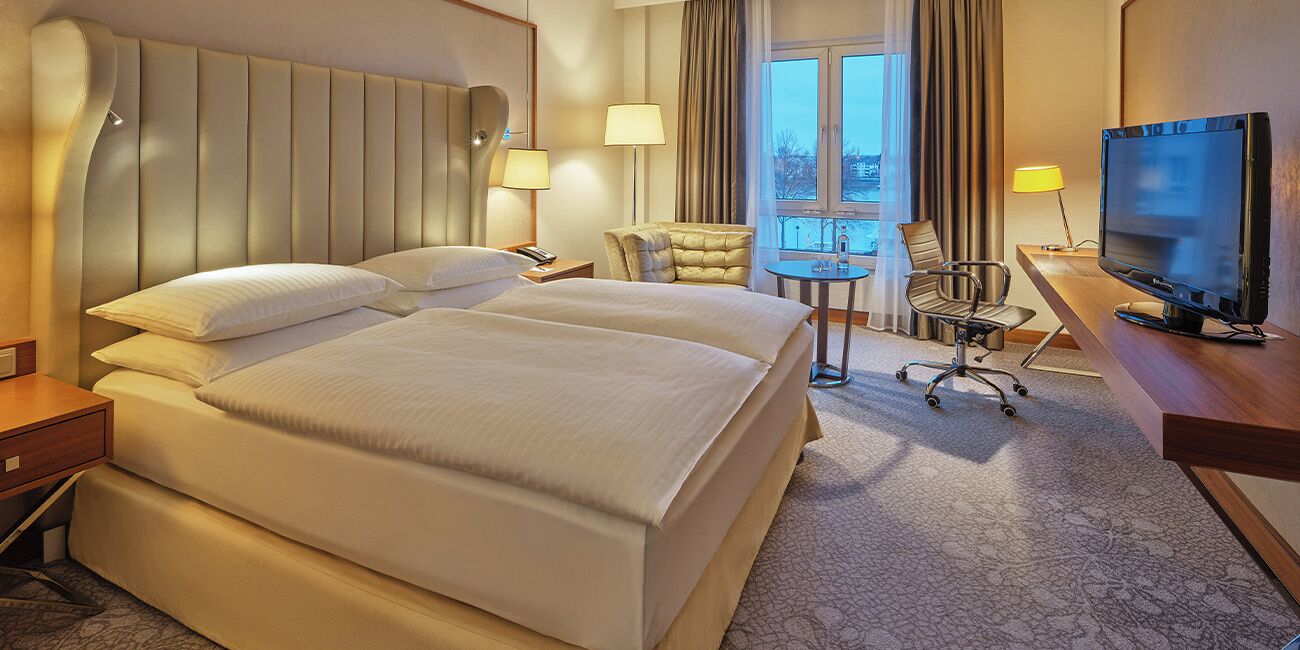 A hotel room with a large bed, a desk with a television, a chair, and a small table.