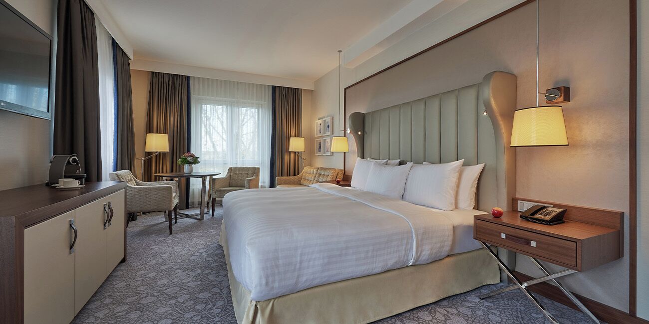 A hotel room with a large bed, a desk, a chair, a TV, and a bedside table with a lamp.