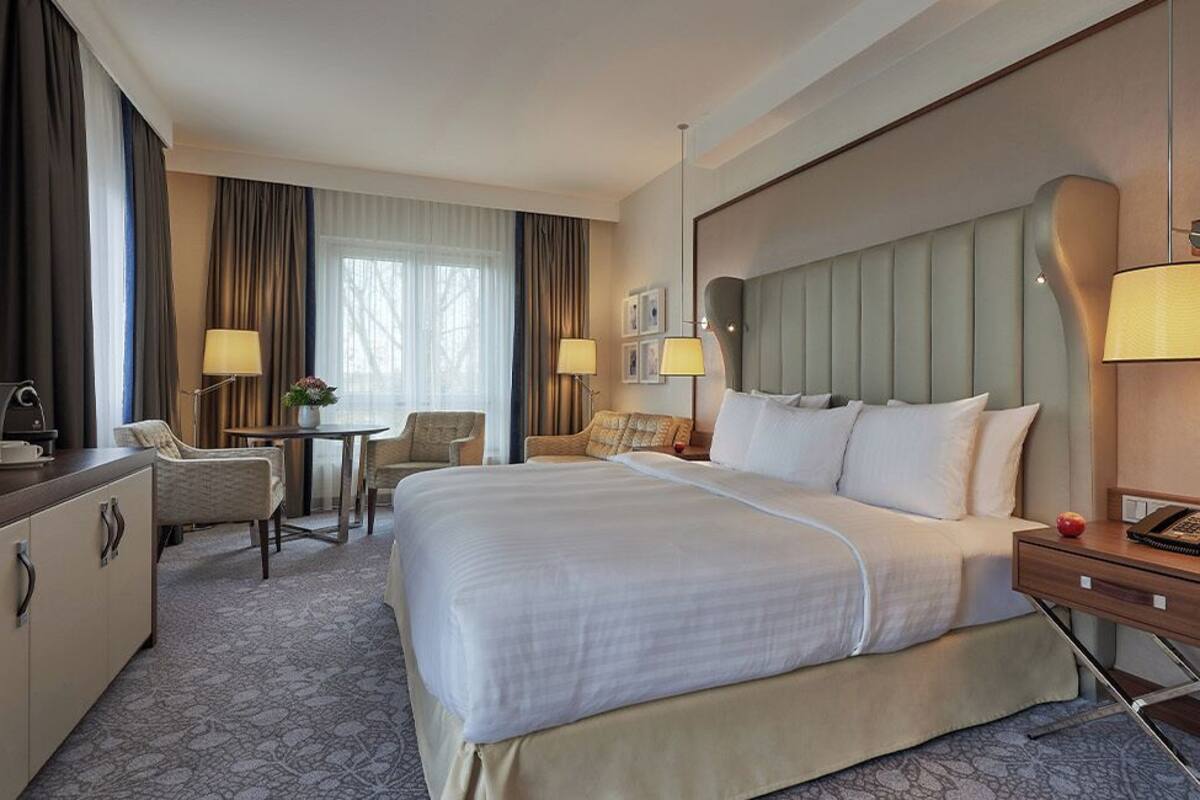 A hotel room with a large bed, a desk, a chair, a TV, and a bedside table with a lamp.
