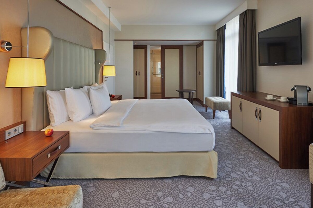A hotel room with a large bed, bedside tables, a desk, a chair, a television, and a wardrobe.