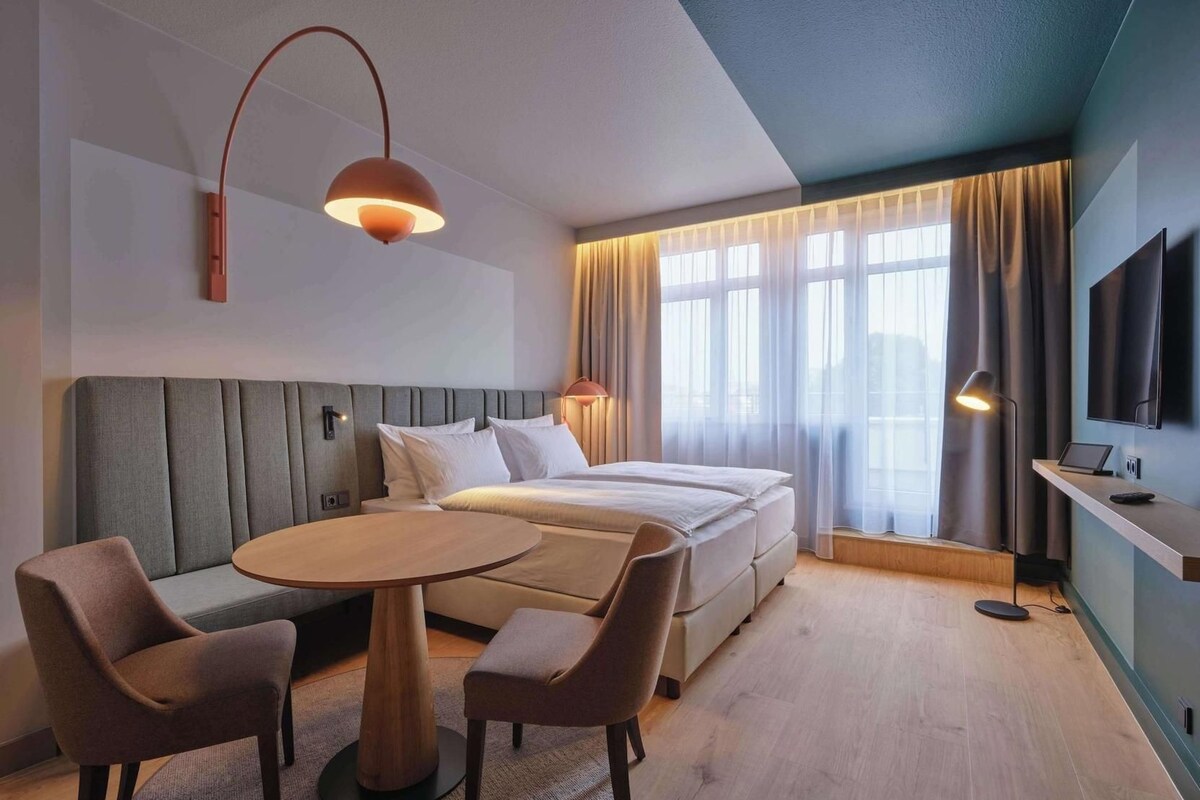 A hotel room with a bed, a round table, two chairs, a TV, and a window with curtains.