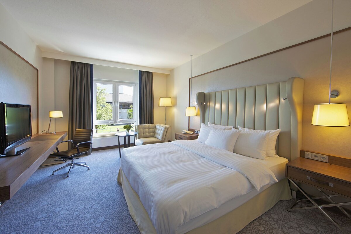 A hotel room with a bed, desk, chair, TV, lamps, and a window with curtains.