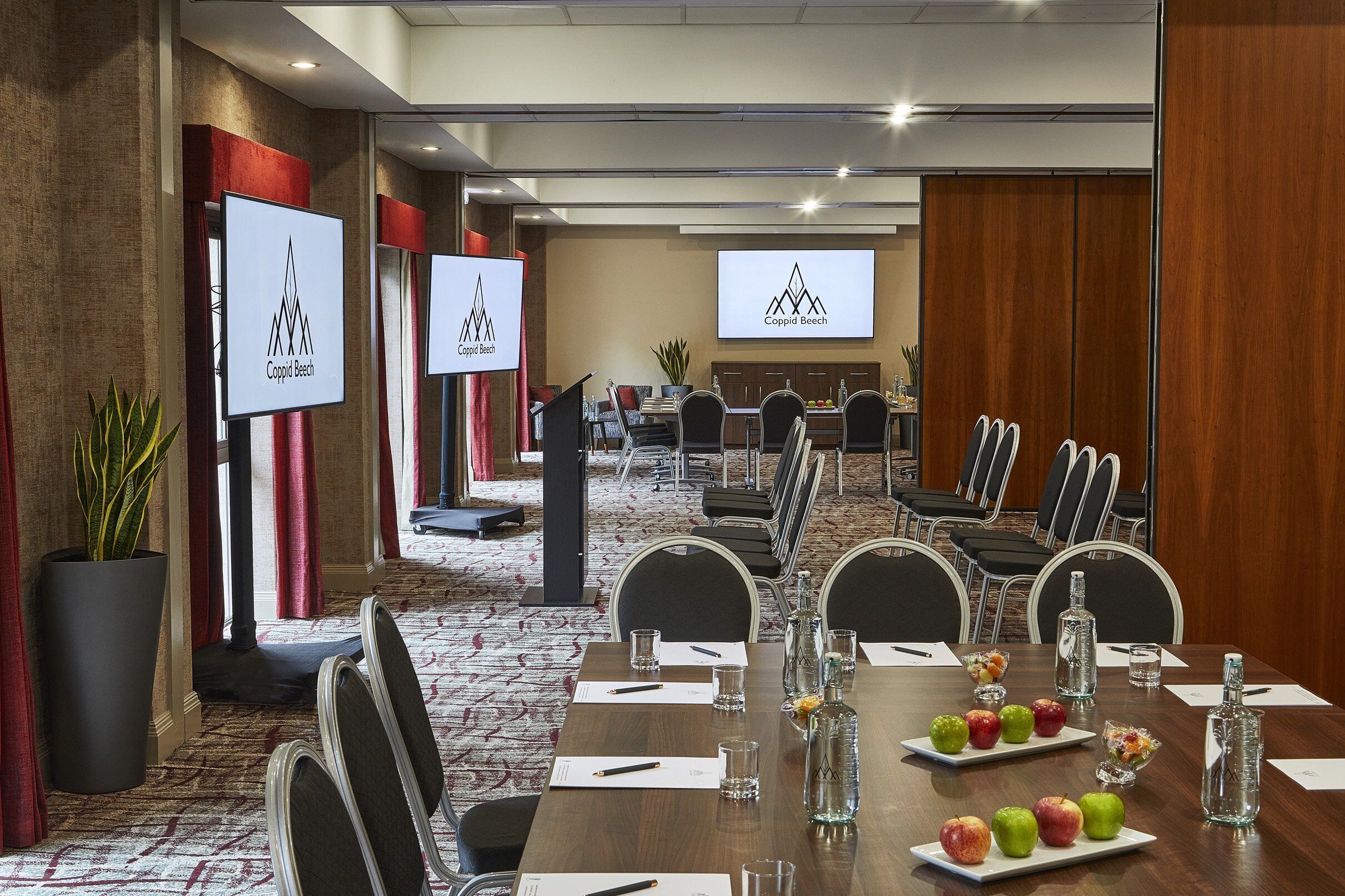 A conference room with a long table, chairs, and a podium with a screen displaying the Coppins Beech logo.