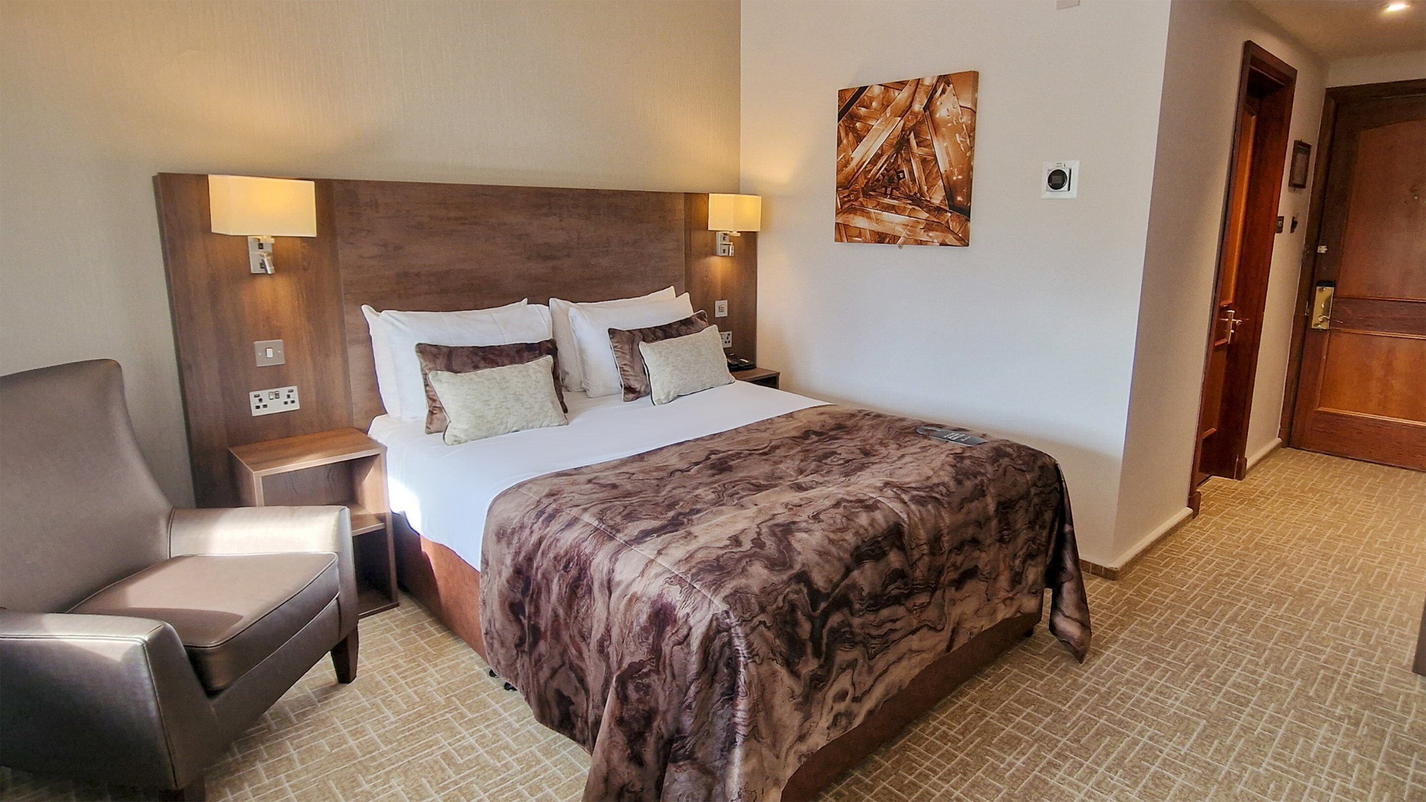 Una habitación de hotel con cama, sillón, mesita de noche, lámpara de pared y una puerta.