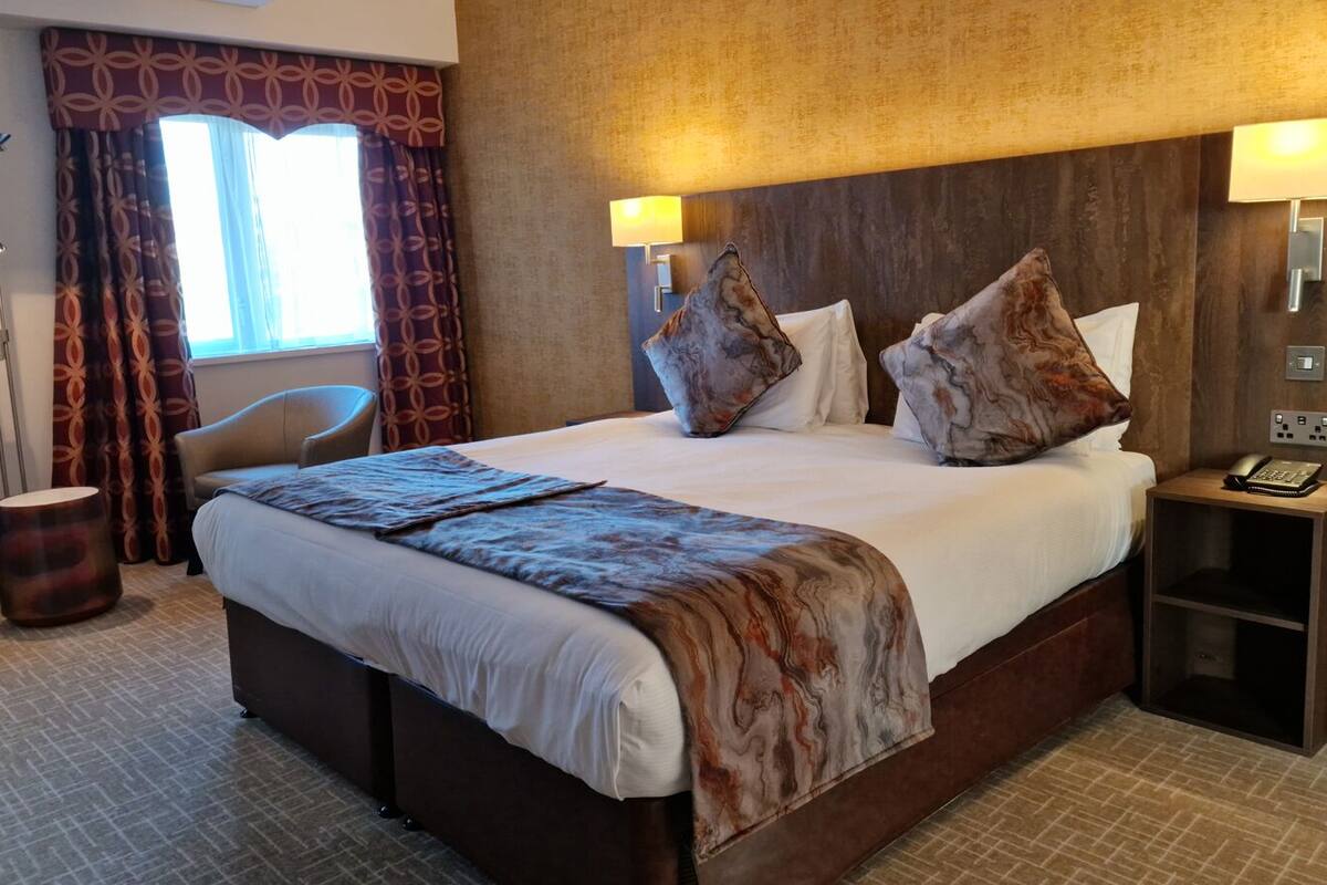 A hotel room with a large bed, two bedside lamps, a telephone on a nightstand, a chair, and a window with curtains.