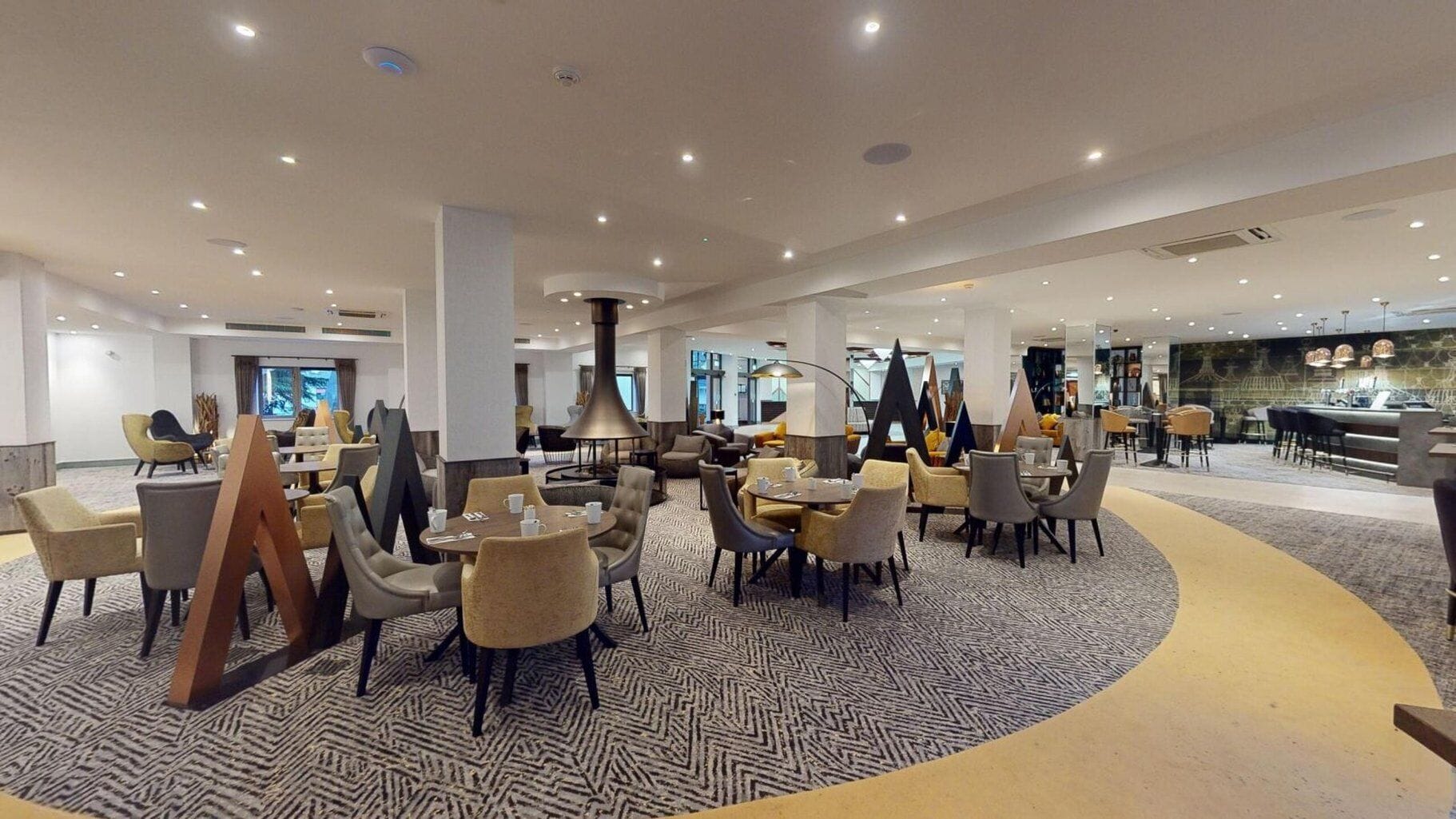 Un hall d’hôtel spacieux, doté de plusieurs espaces de détente, notamment des tables et des chaises rondes, ainsi qu’un espace central agrémenté d’un tapis à motifs.