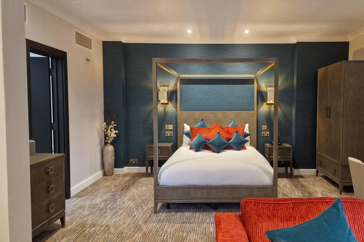 A modern hotel room with a four-poster bed, a large mirror, a wooden wardrobe, and a red and blue sofa.