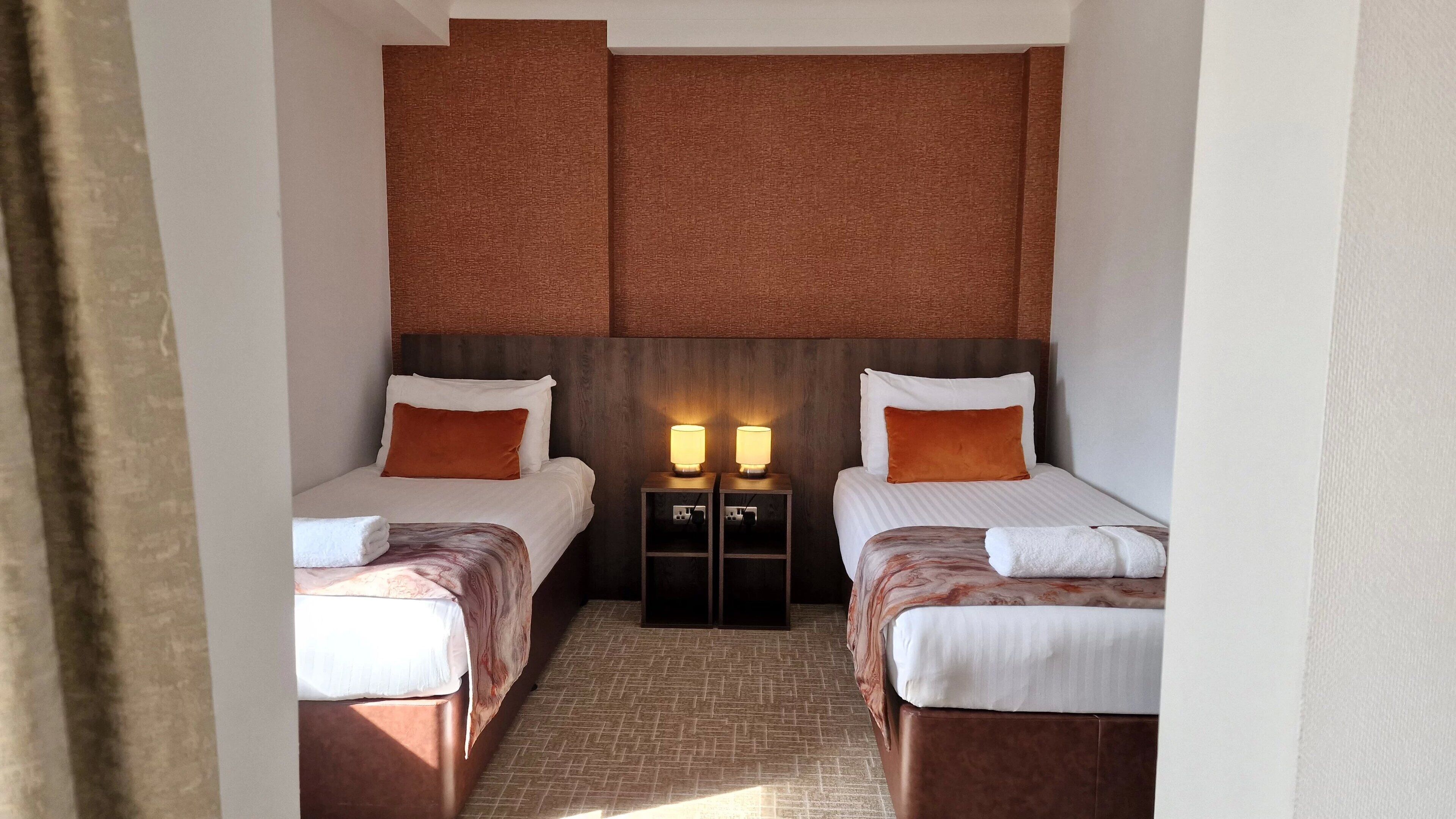 A hotel room with two single beds, a wooden headboard, a nightstand with lamps, and a tiled floor.