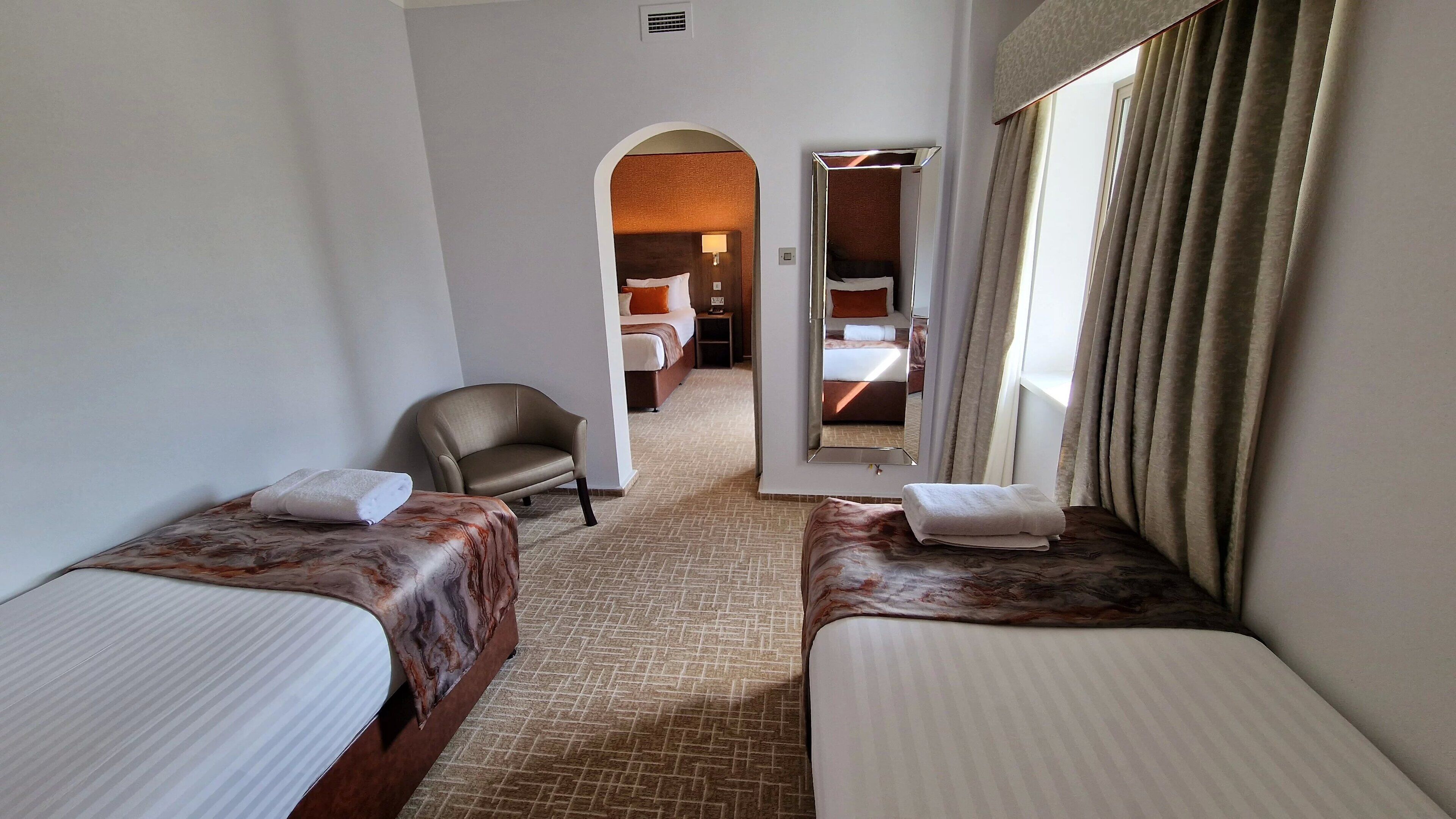 A hotel room with two single beds, a chair, a mirror, and a view of another room.