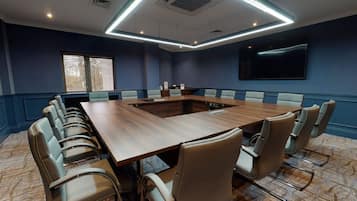 Uma sala de conferência com uma mesa comprida, várias cadeiras, uma tela grande e uma janela com persianas.
