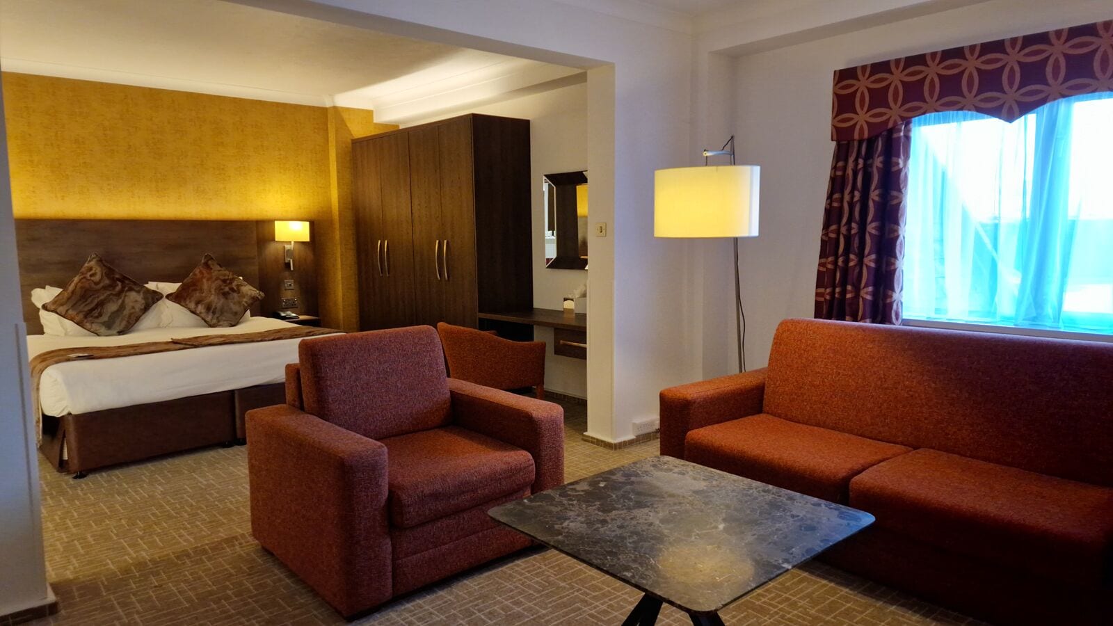 A hotel room with a bed, two armchairs, a sofa, a small table, a lamp, and a window with curtains.