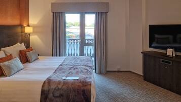 A hotel room with a large bed, a TV, a balcony with curtains, and a wooden headboard.