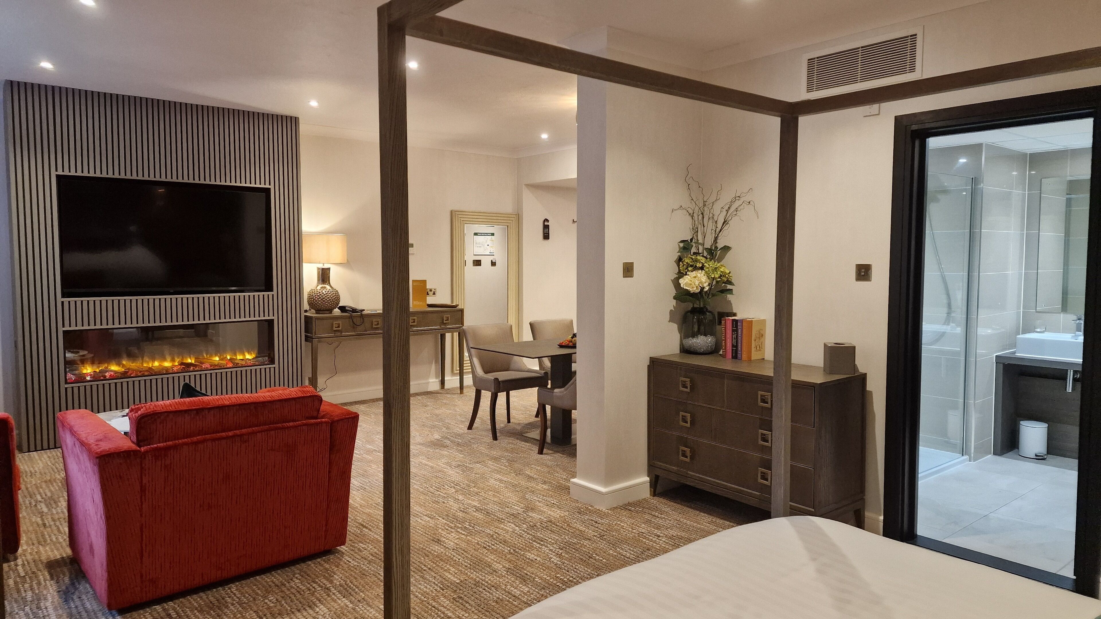 A modern hotel room with a flat-screen TV, a fireplace, a red sofa, a dining area, and a bathroom visible through a glass door.