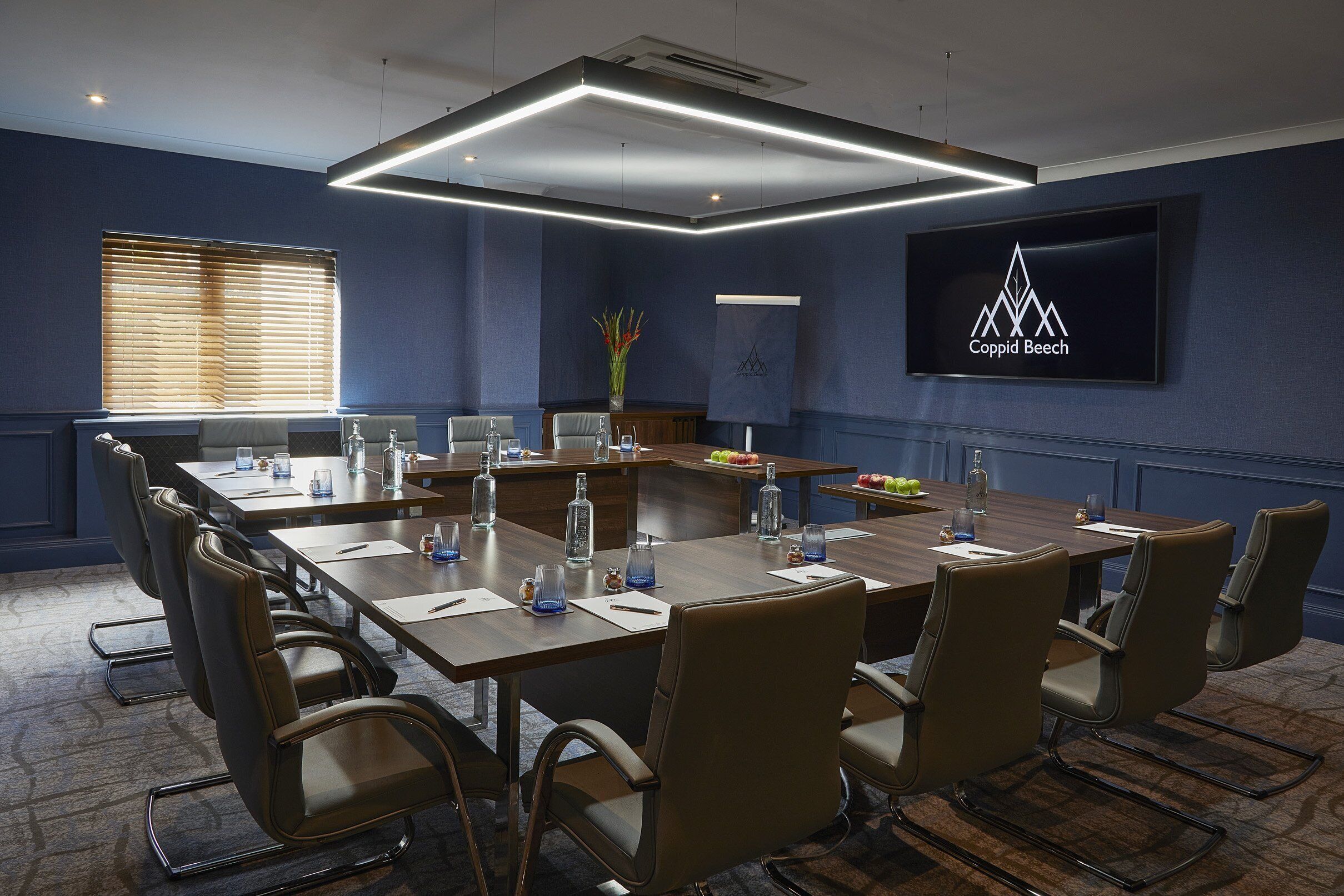 Un salón de conferencias con una larga mesa, sillas y una pantalla mostrando el logotipo de Coppid Beech.