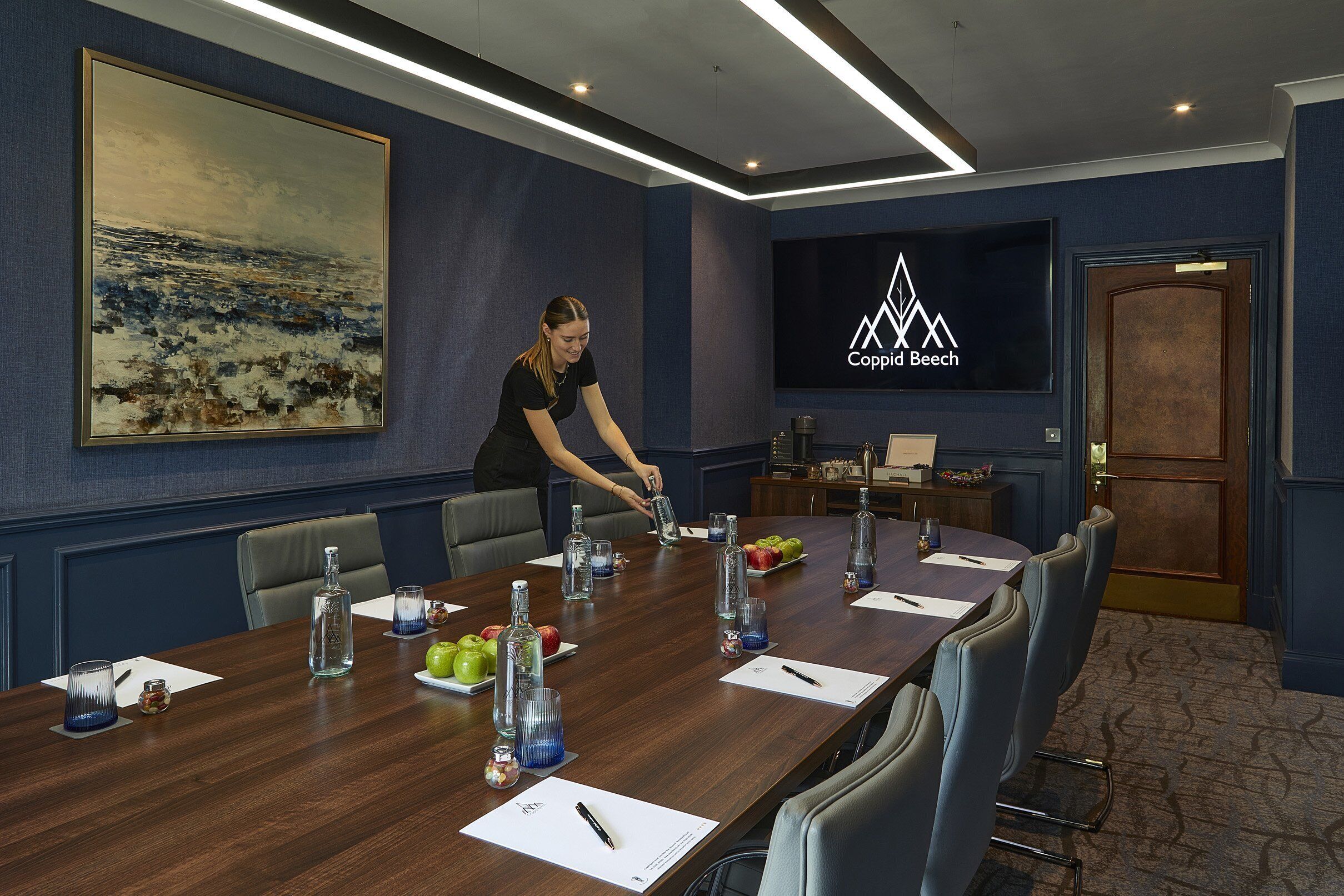 Un salón de conferencias con una mesa larga, sillas, una pantalla de televisión y un cuadro en la pared.