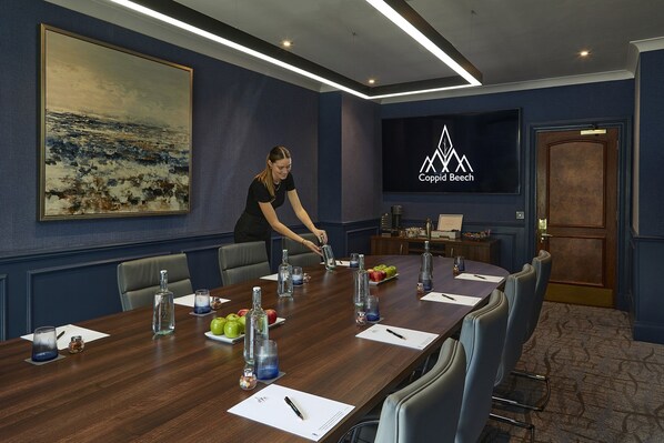 Un salón de conferencias con una mesa larga, sillas, una pantalla de televisión y un cuadro en la pared.