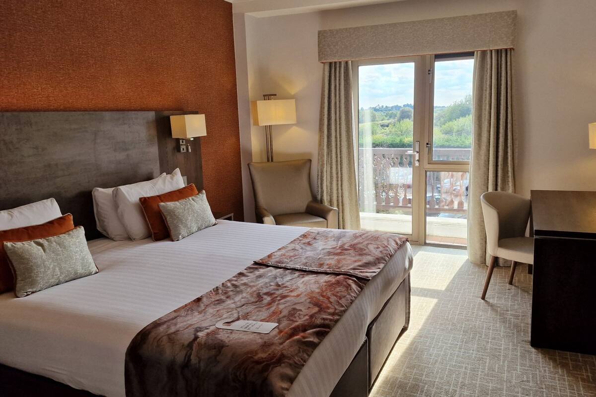 A hotel room with a large bed, two bedside tables, a desk, and a chair. There is a view of a balcony through a sliding door.