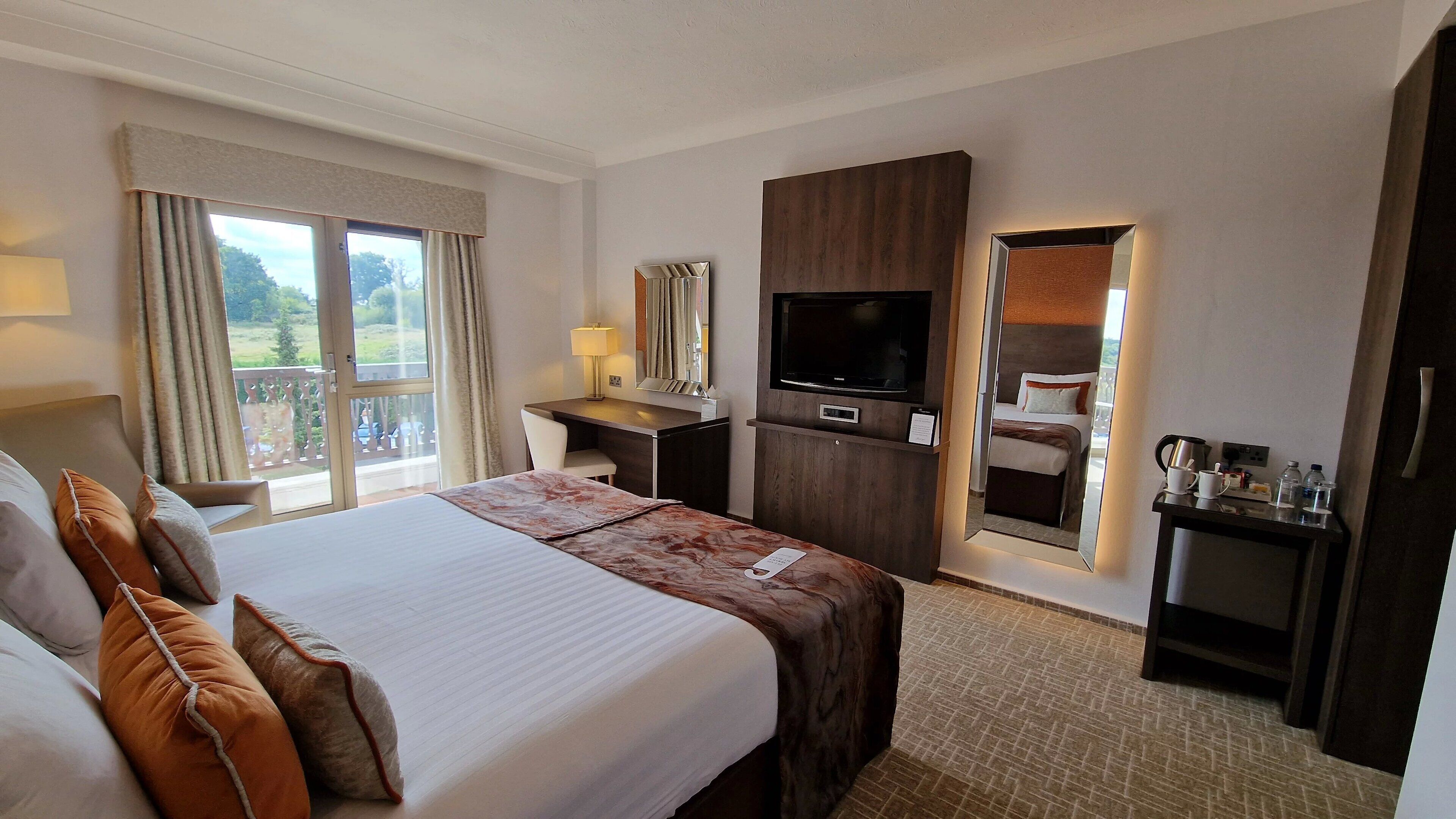 A hotel room with a large bed, a desk with a flat-screen TV, a mirror, and a balcony with curtains.