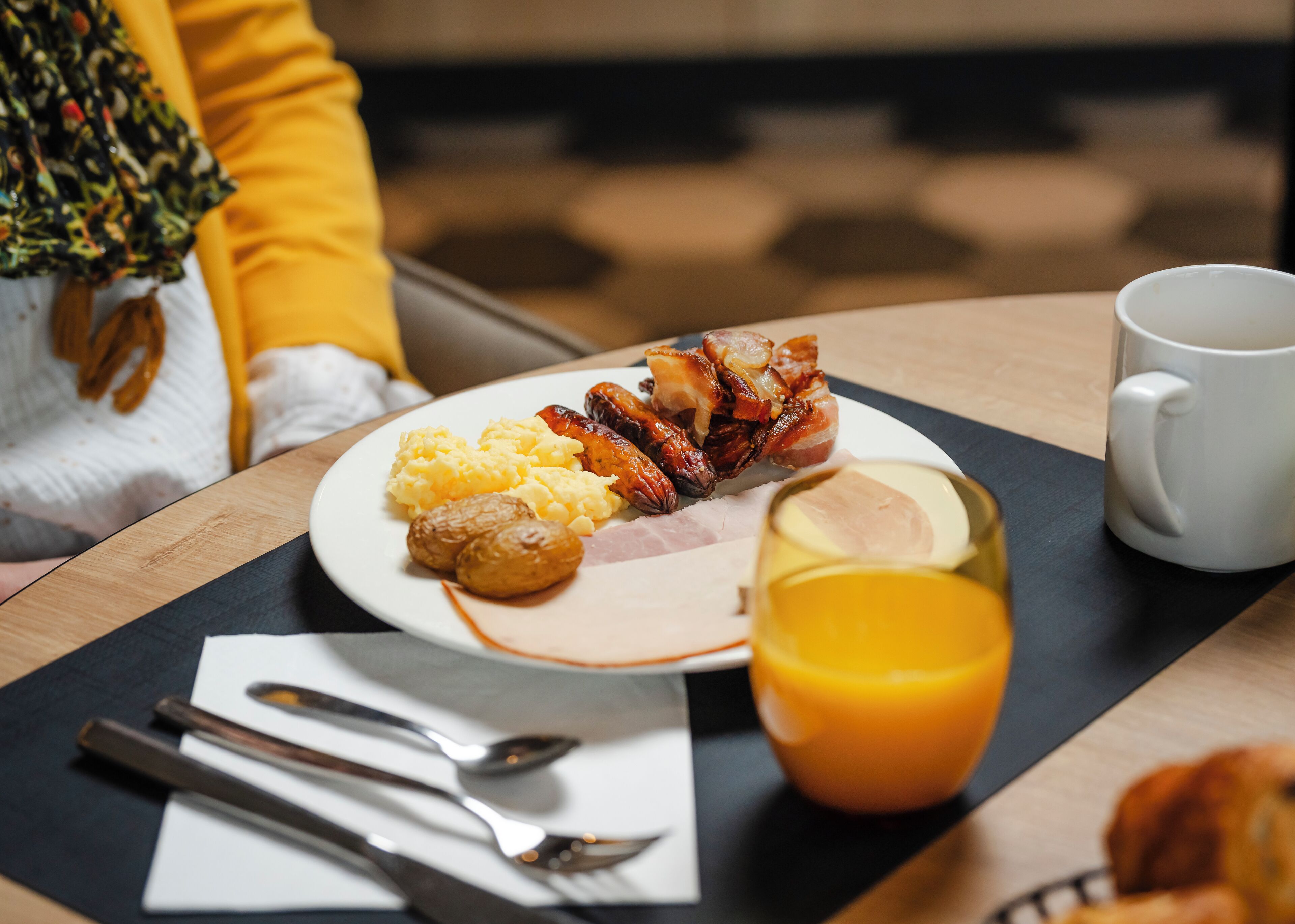 En frukosttallrik med ägg, korv och potatis, ett glas apelsinjuice och en vit mugg på ett träbord.