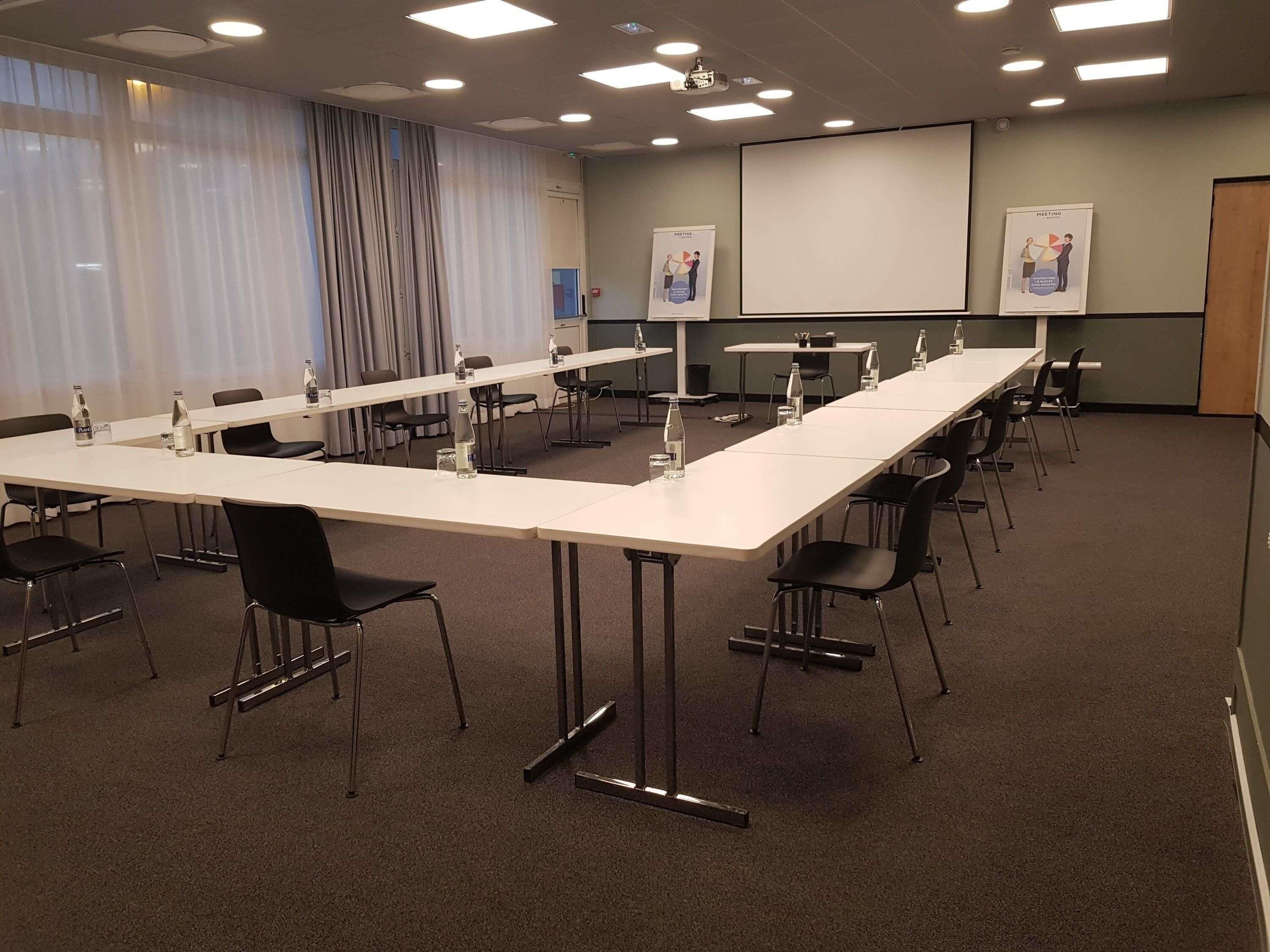 Une salle de conférence avec de longues tables, des chaises, un écran de projection et deux affiches au mur.