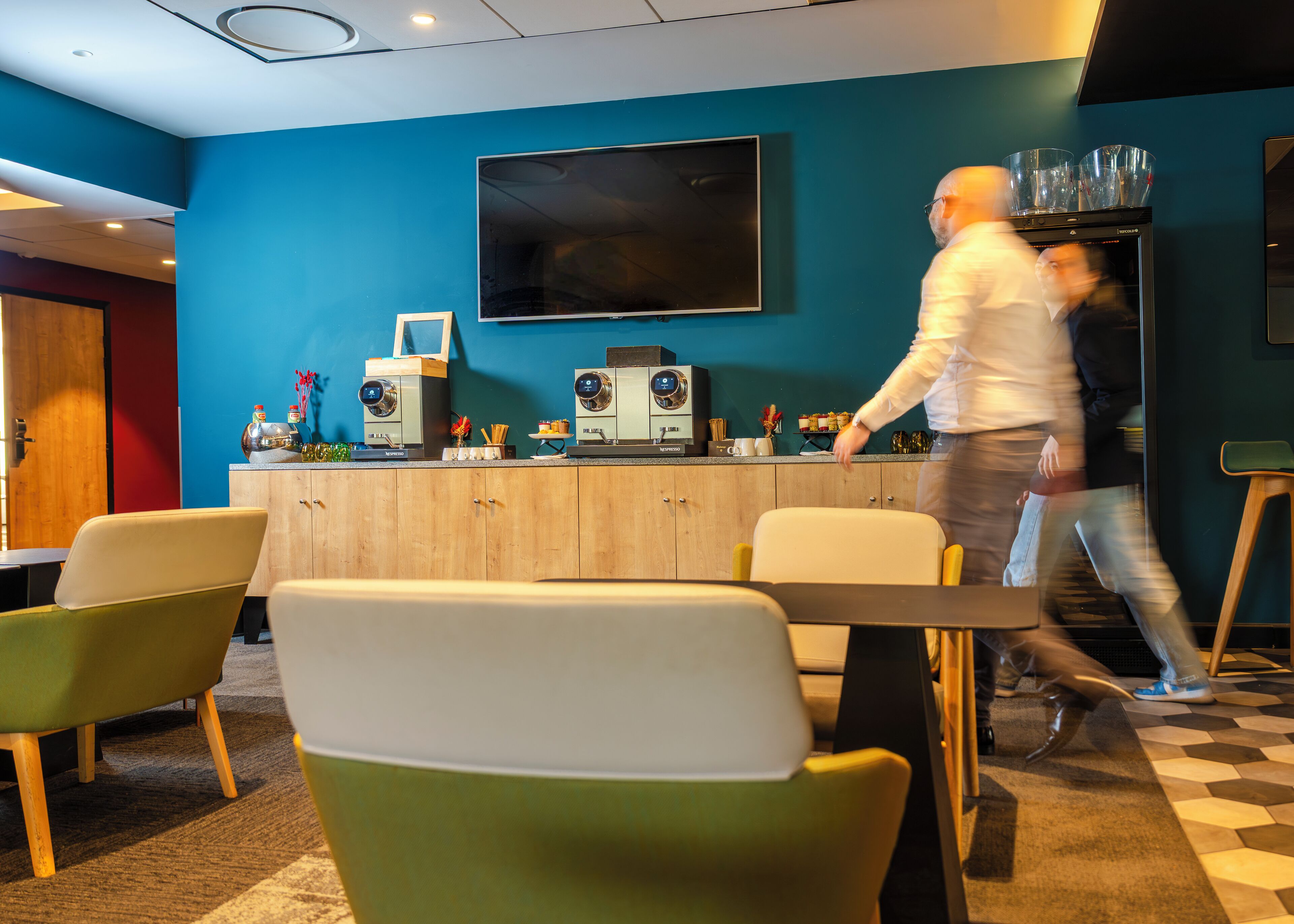 A modern bar area with a flat-screen TV, two espresso machines, and a counter with various items. There are colorful chairs and a checkered floor.