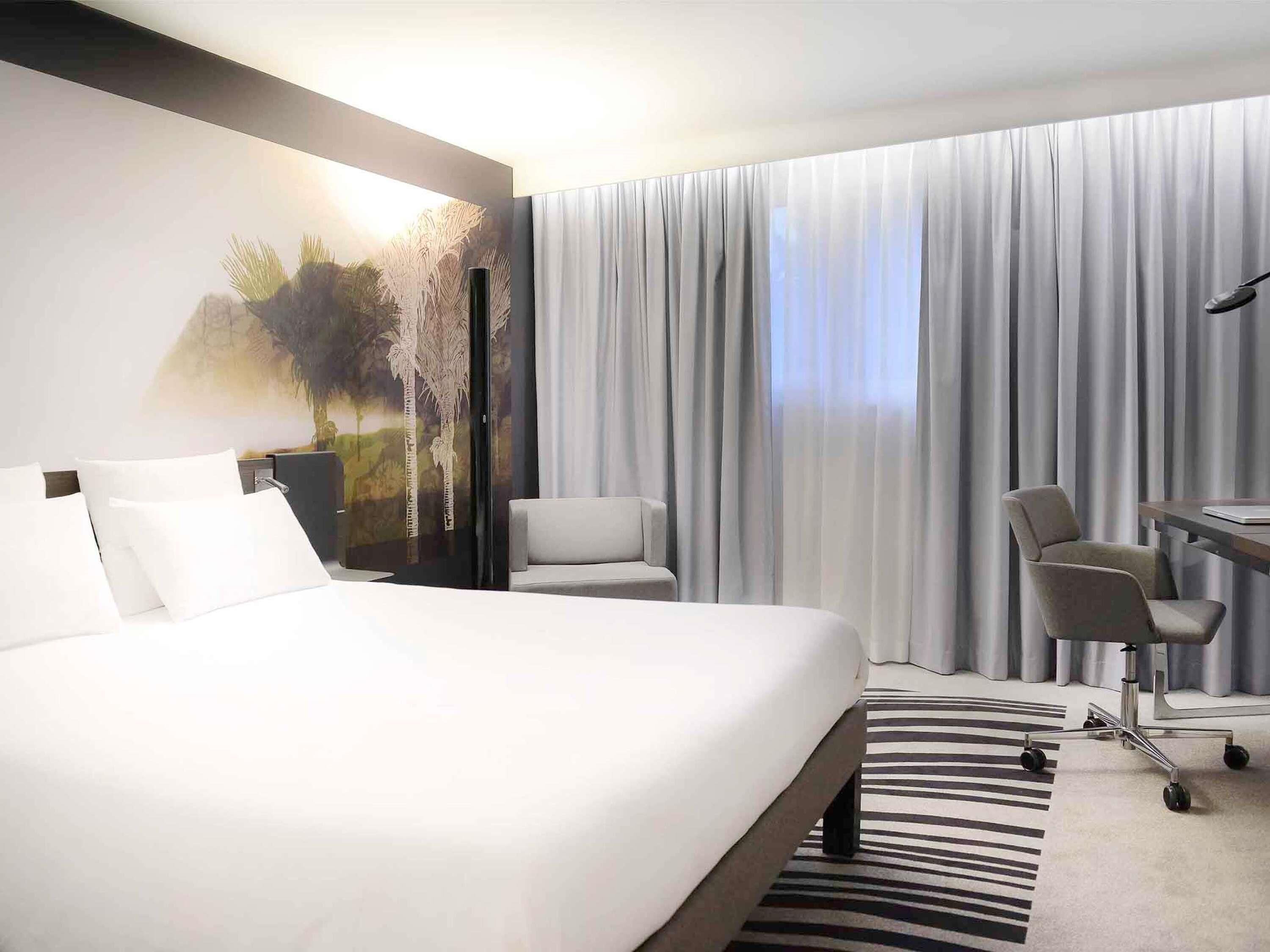 A hotel room with a large bed, a desk with a chair, a view of the outdoors through sheer curtains, and a painting on the wall.