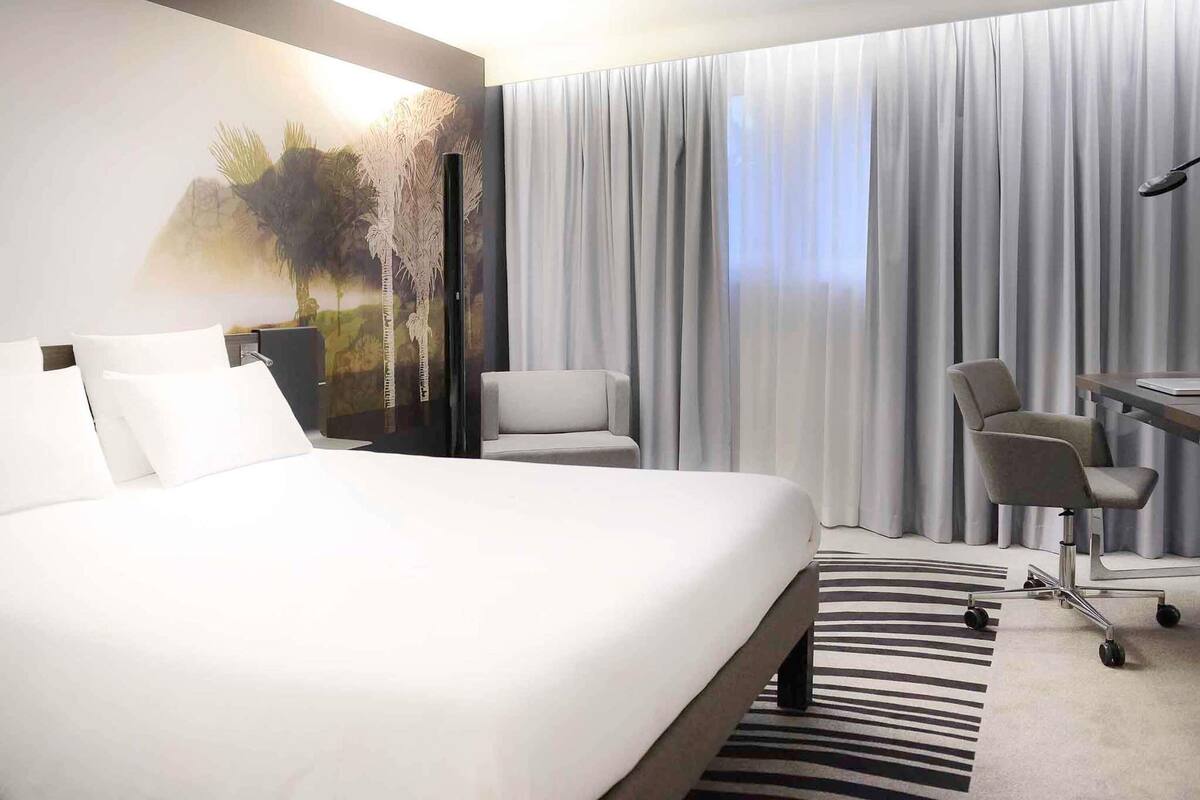 A hotel room with a large bed, a desk with a chair, a view of the outdoors through sheer curtains, and a painting on the wall.